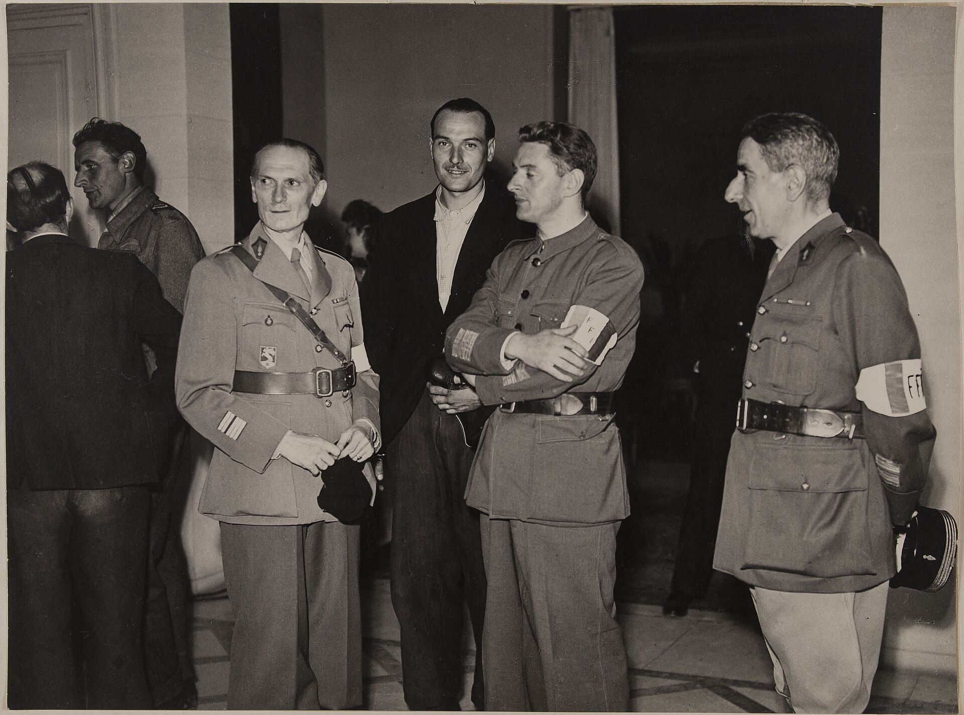 photographie de 1944 en noir et blanc avec quatre hommes dans un salon, trois en uniforme avec le brassard ffi et un en veste civile, a la reception de l etat-major ffi