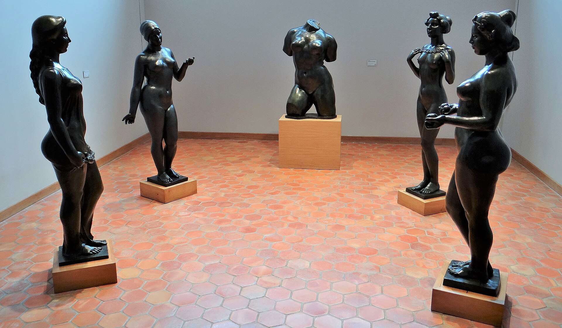 une salle du musee maillol avec cinq bronzes de maillol sur des socles de bois clair, des murs blancs et un sol de tomettes hexagonales rouges