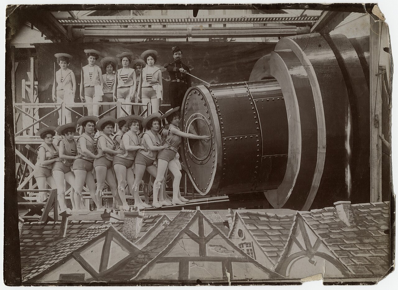 fotografia di scena del 1902 con una fila di giovani donne in costume alla marinara che spingono la capsula nella bocca del cannone, un uomo in divisa su una passerella e il fondale dipinto dei tetti in basso