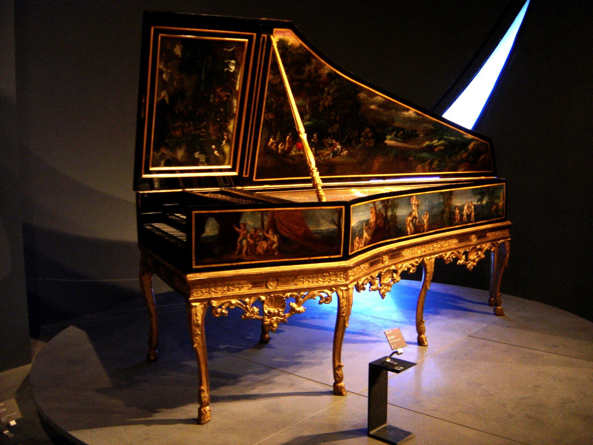 clavicembalo con la cassa nera filettata d oro su un sostegno rococo dorato, il coperchio alzato dipinto con un paesaggio popolato di figure e animali, illuminato nella sala buia del museo