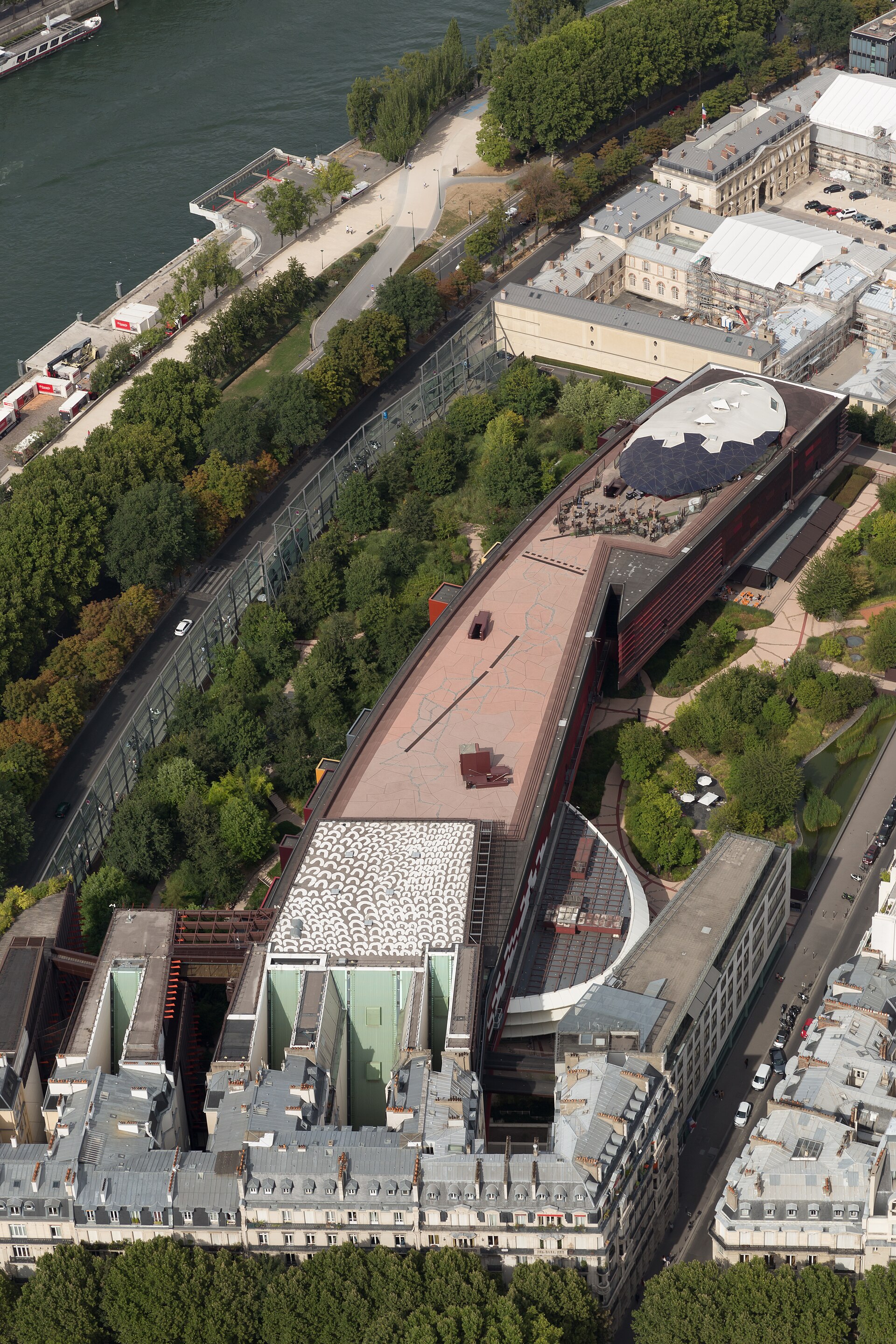 le musee du quai branly vu du haut de la tour eiffel, avec le long batiment courbe pose parmi les arbres du jardin, les boites en saillie sur la facade et la seine en haut a gauche