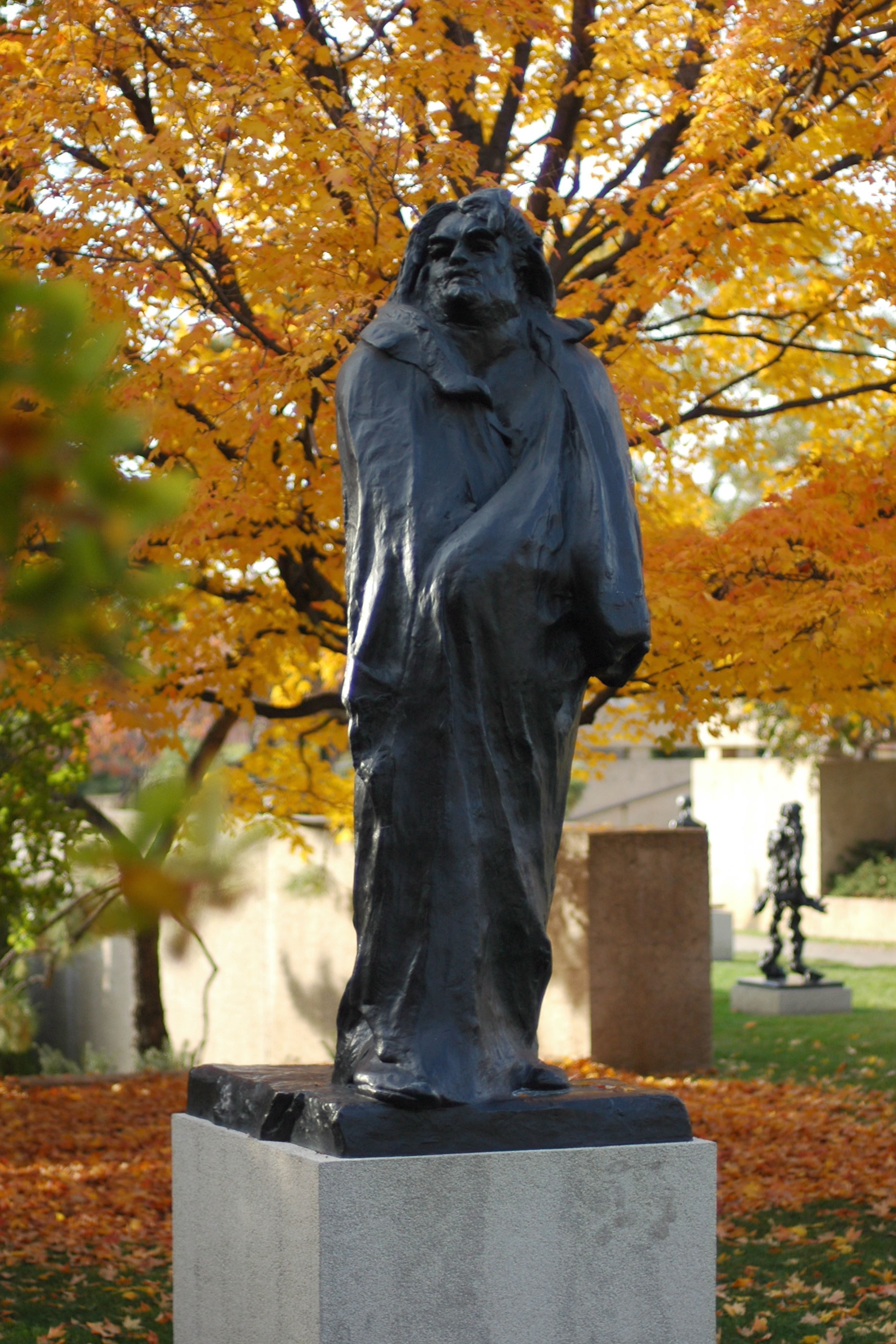 le monument a balzac de rodin dans le jardin du musee en automne, une figure de bronze sombre enveloppee dans une longue robe de chambre, la tete haute, avec des arbres jaunes derriere