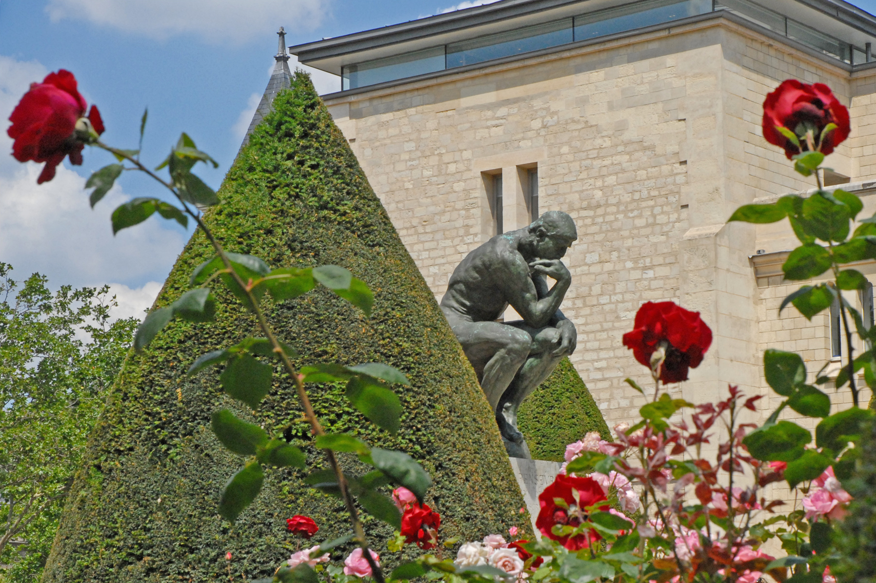 le penseur de rodin sur son socle dans le jardin du musee, parmi les roses rouges et les buis tailles en cone, avec la facade de l hotel biron au fond