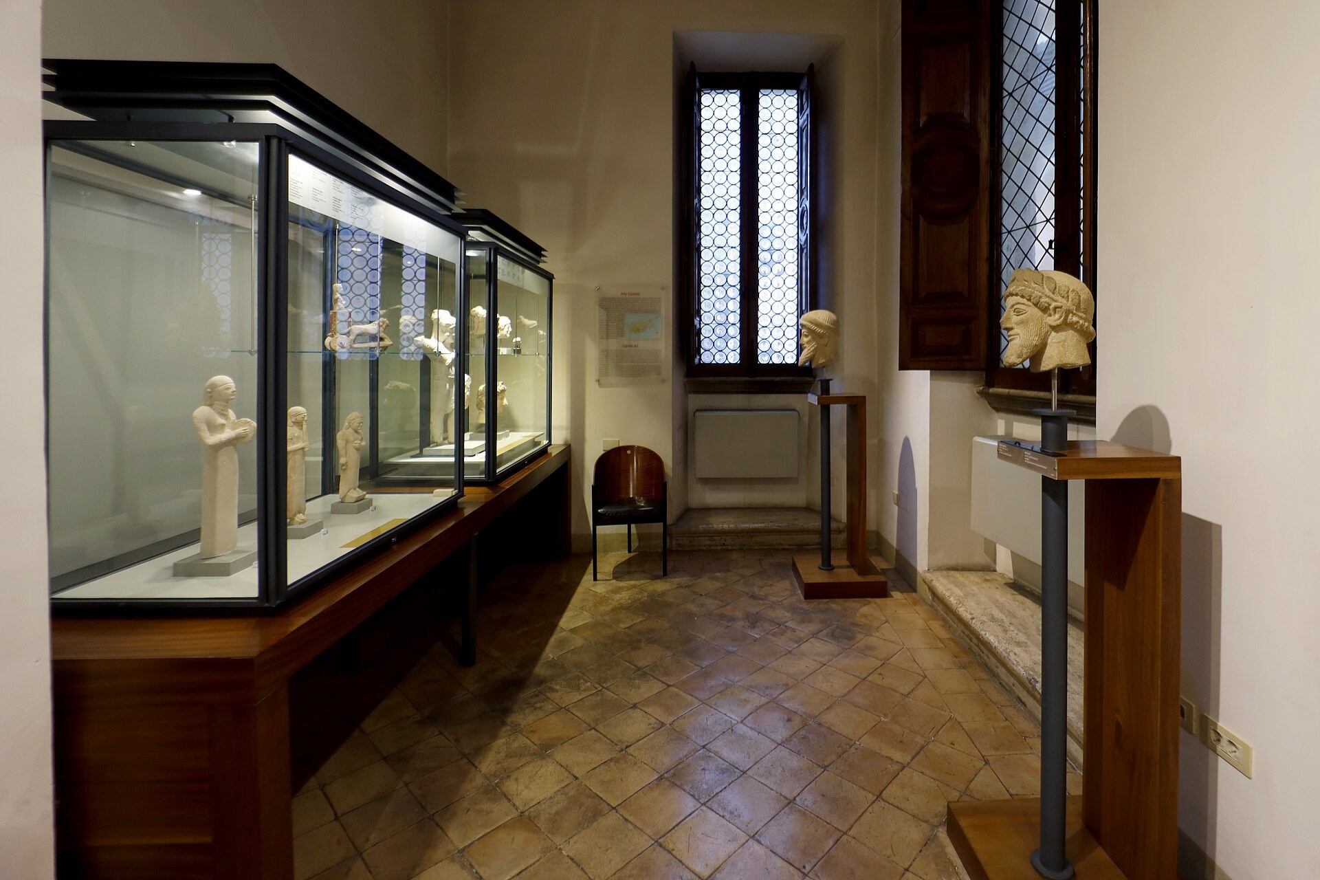 la sala del arte chipriota del museo barracco, con la larga vitrina iluminada a la izquierda llena de estatuillas de caliza, dos cabezas barbudas sobre columnitas metalicas a la derecha y el suelo de barro cocido
