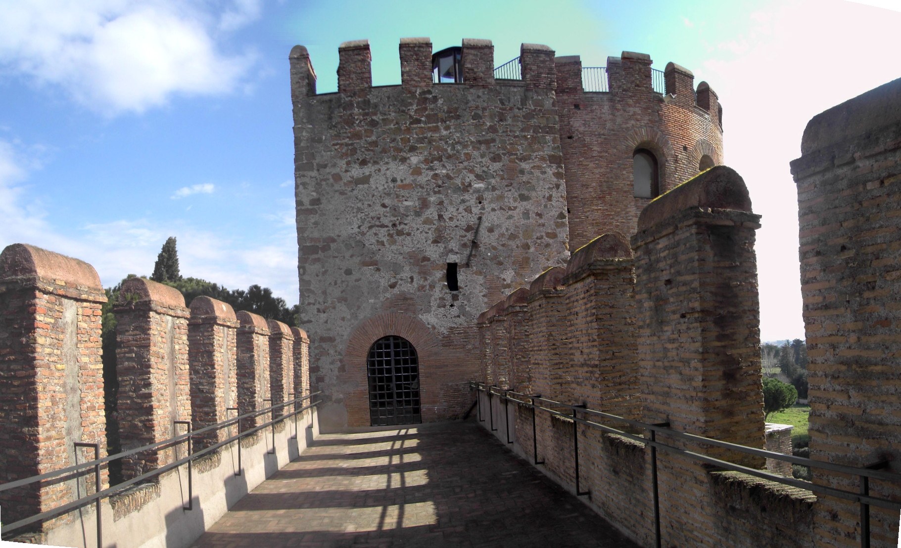 el tramo descubierto del paso de ronda en porta san sebastiano, con las almenas de ladrillo a la izquierda, la barandilla moderna a los dos lados y al fondo una torre cuadrada con la puerta de reja
