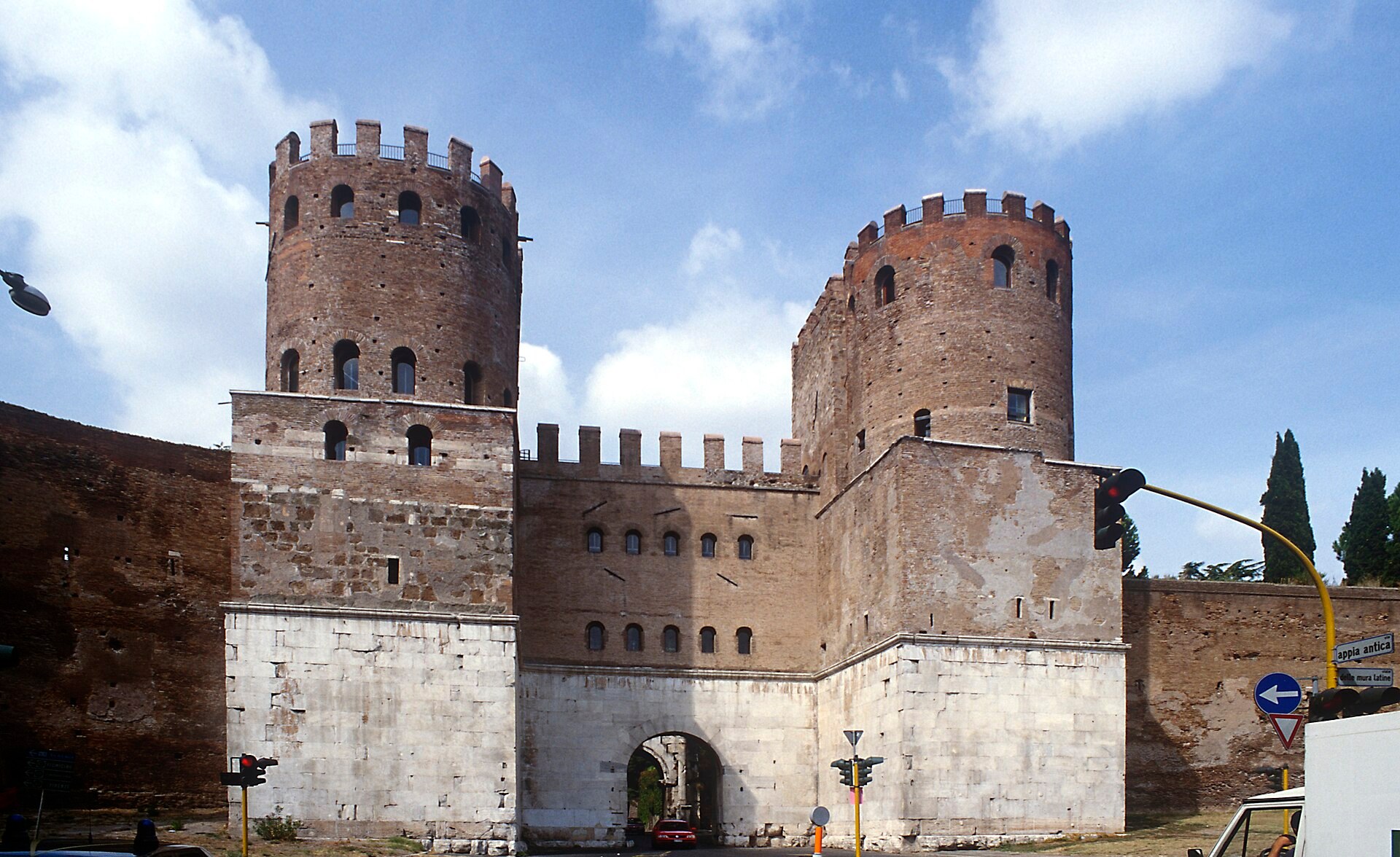porta san sebastiano vista desde fuera, con las dos torres redondas almenadas de ladrillo, el basamento revestido de marmol blanco y el arco abierto en el centro