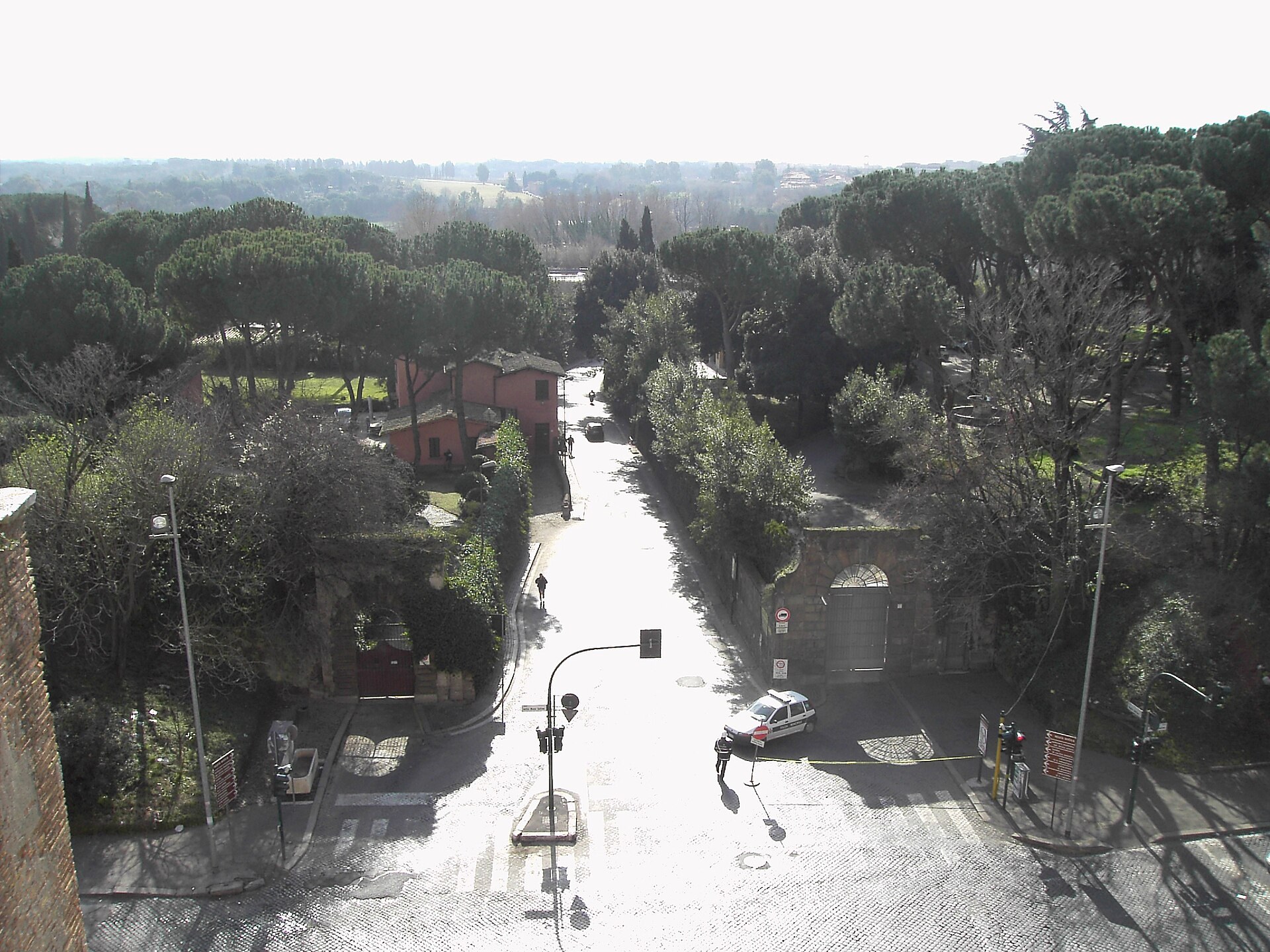 la via appia vista desde lo alto de porta san sebastiano, la calle recta que se aleja entre los muros de los jardines y los pinos pineos, con una casa roja a la izquierda
