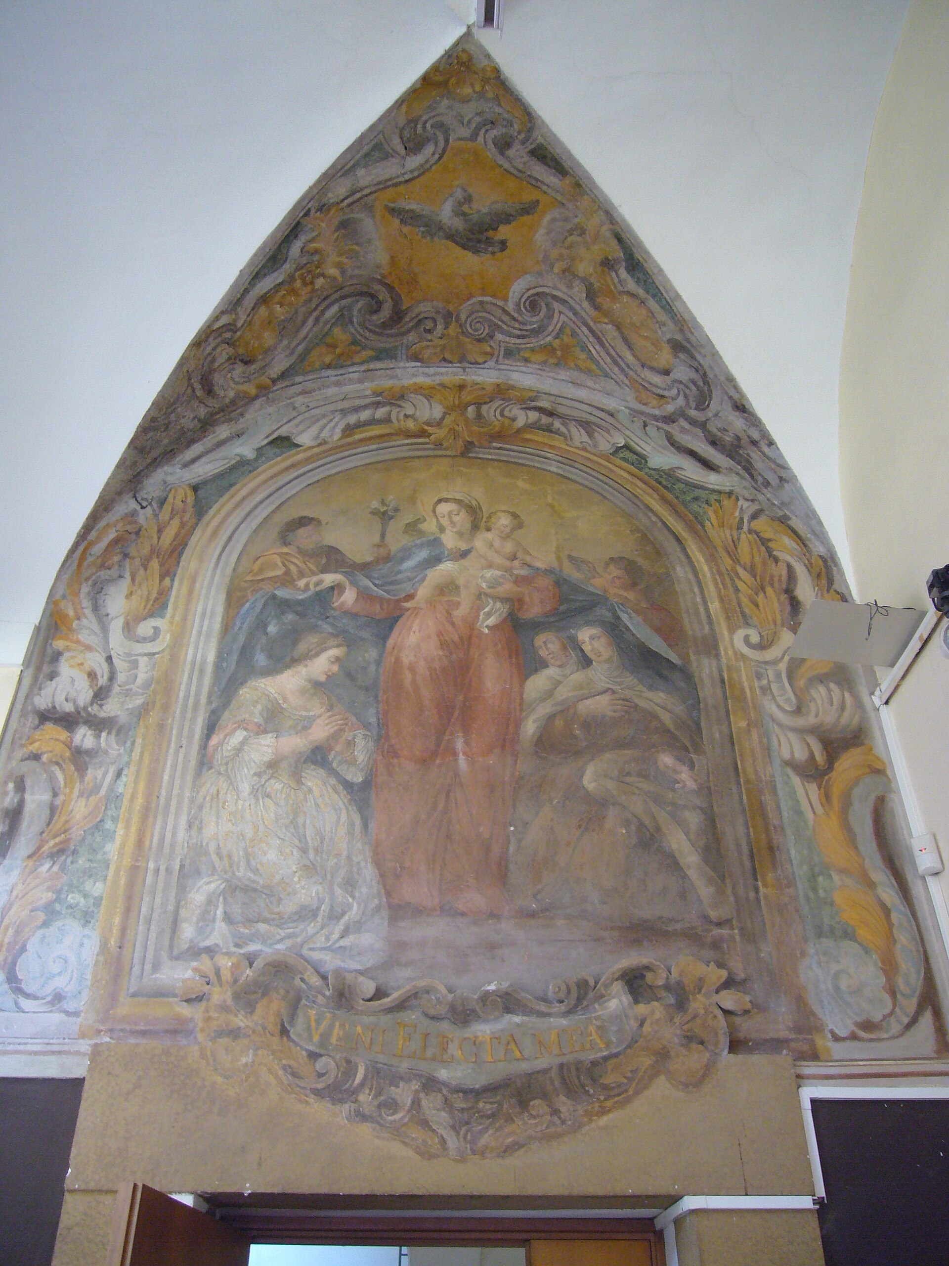 la lunette a fresque de l ancien couvent de sant egidio, avec la vierge a l enfant au centre, une jeune femme agenouillee en blanc a gauche, deux carmelites voilees a droite, la colombe du saint esprit dans le tympan et le cartouche veni electa mea en bas