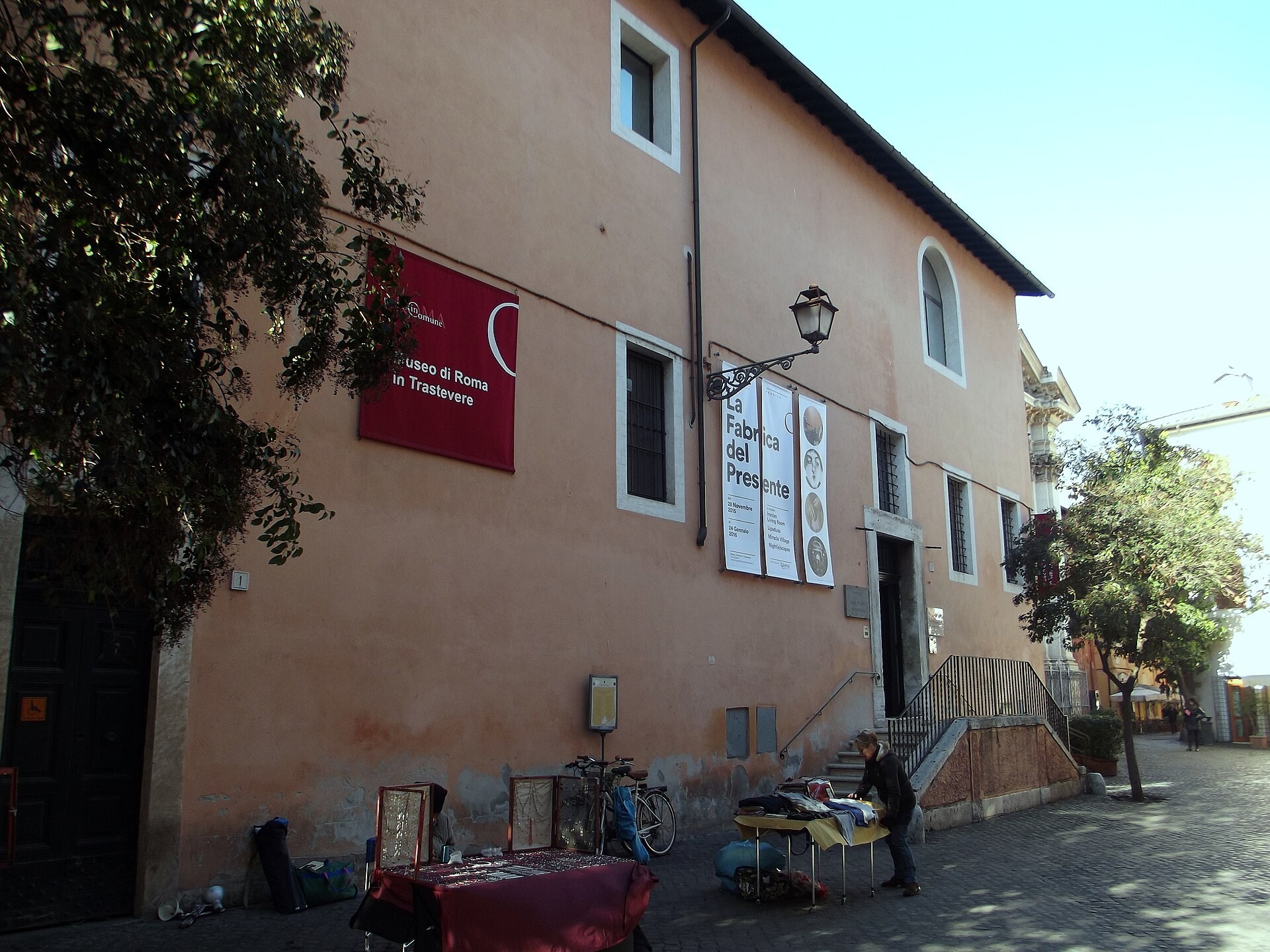 le mur exterieur rose du musee de rome au trastevere avec l enseigne du musee, des etals de tableaux appuyes devant et des arbres a gauche