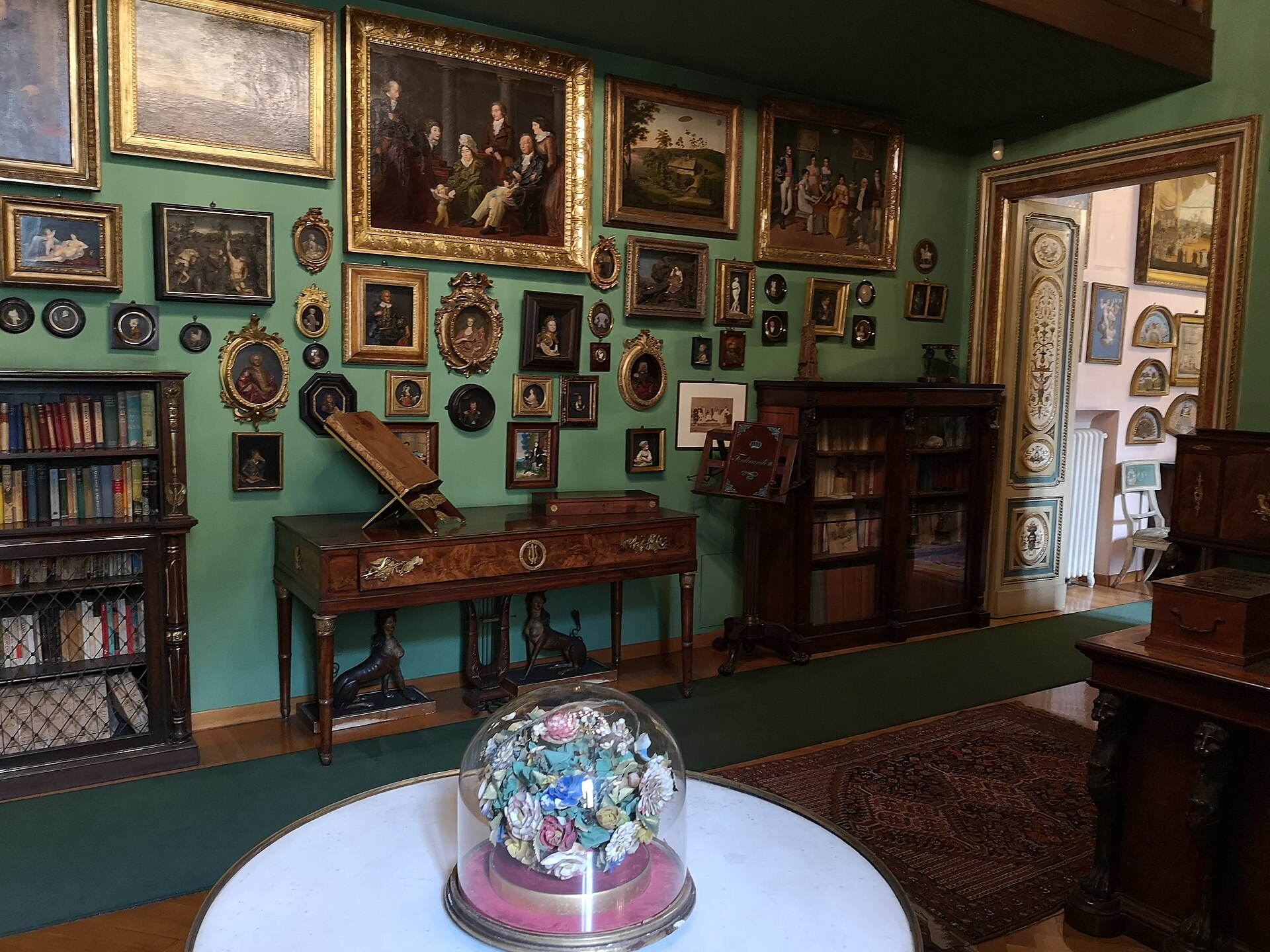 lo studio della casa museo di mario praz a palazzo primoli, con le pareti verdi coperte di quadri e ritratti, i mobili impero e una campana di vetro sul tavolo