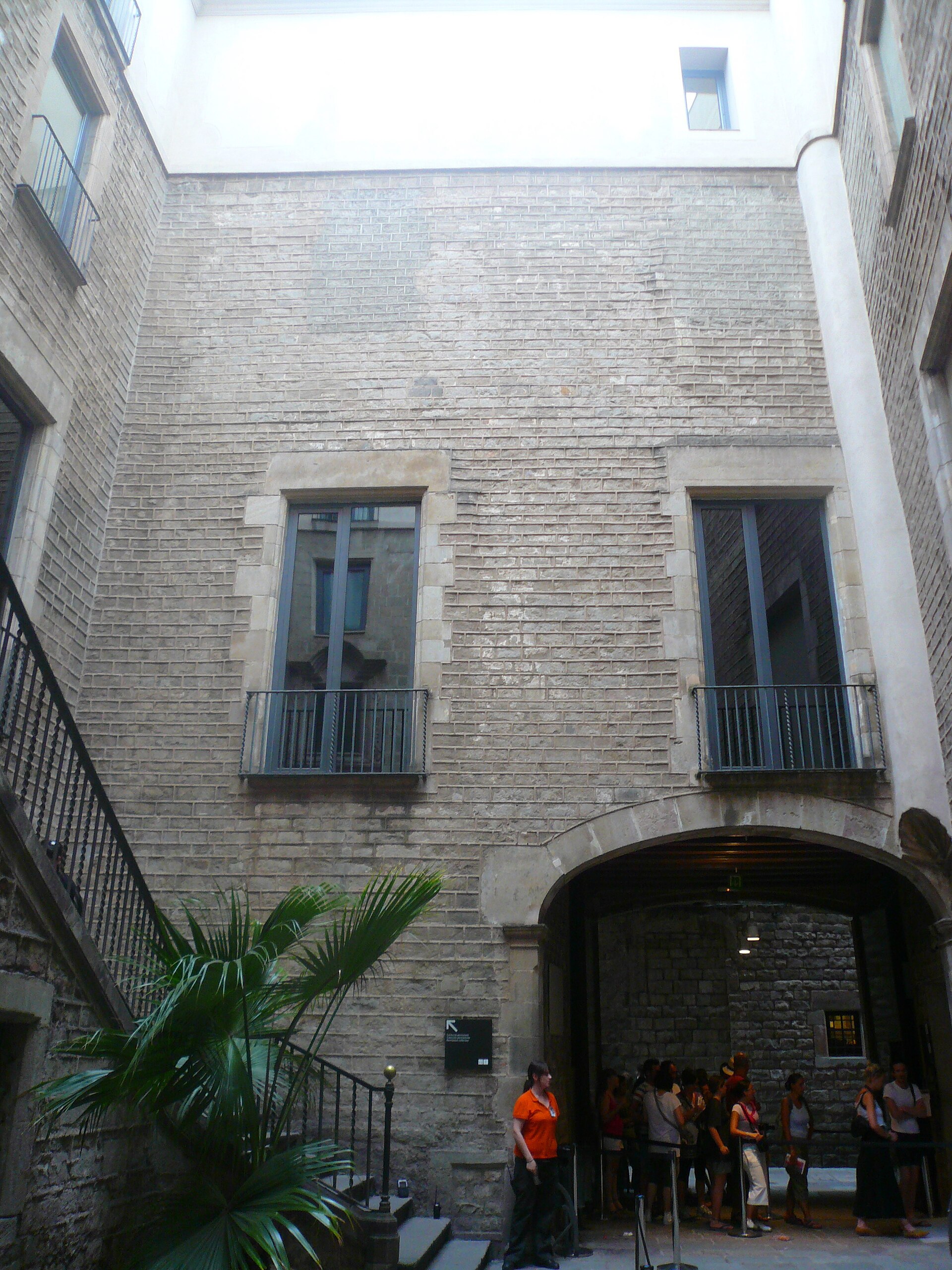 il cortile del palau meca con le pareti di pietra, le finestre con le ringhiere, una scala laterale e un gruppo di visitatori sotto l’arco d’ingresso