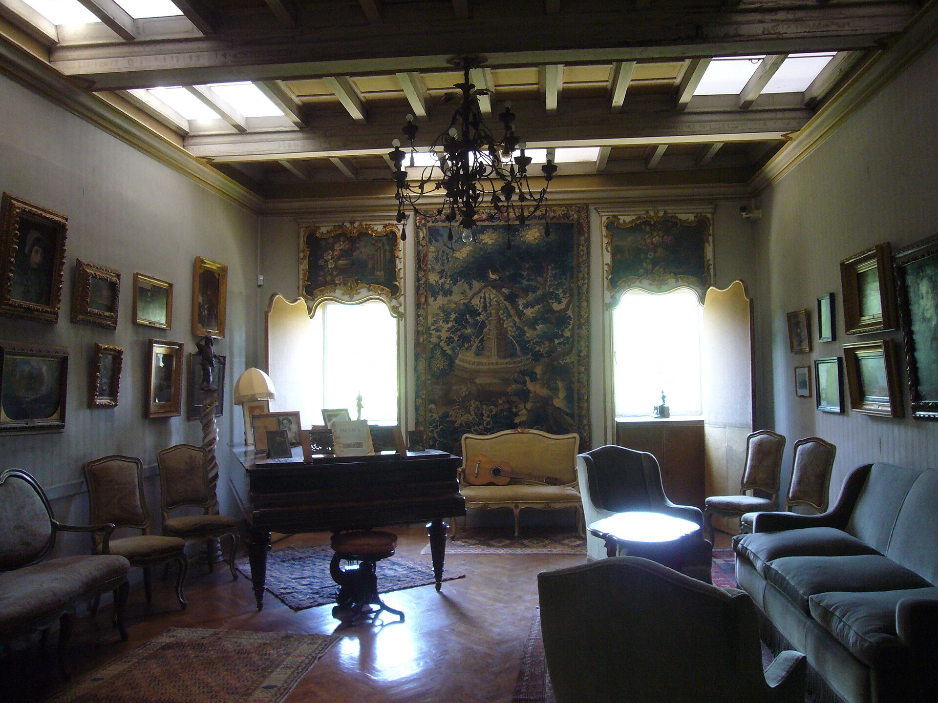el salon del apartamento privado de canonica, con el piano de cola y las partituras abiertas, una guitarra apoyada en el sofa, el gran tapiz de verdura en la pared del fondo, los cuadros colgados muy juntos y la lampara de hierro forjado