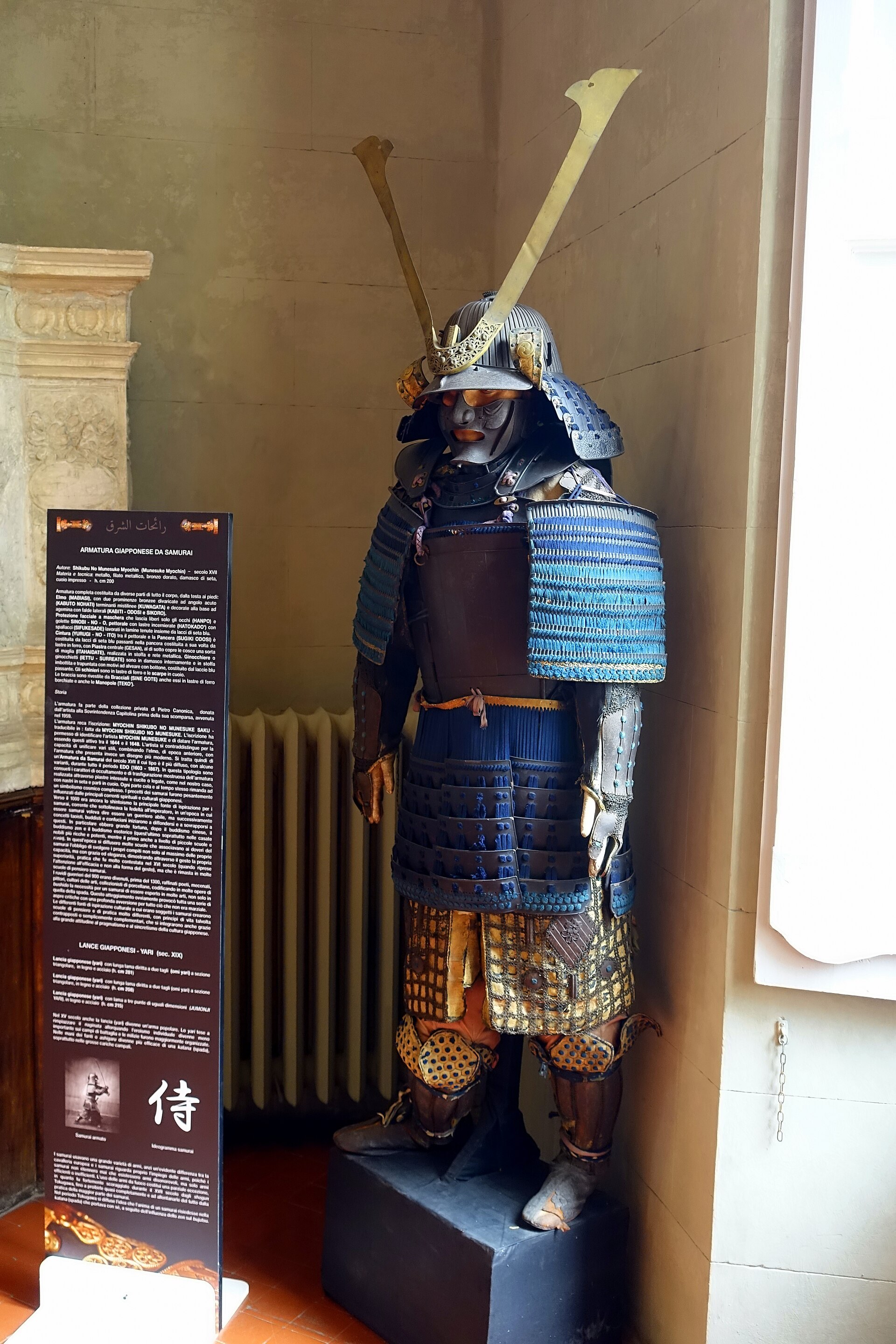 la armadura de samurai expuesta sobre una peana negra, con el yelmo, la mascara facial con bigote, las placas atadas de azul, dos hojas de lanza montadas detras y el panel explicativo a la izquierda