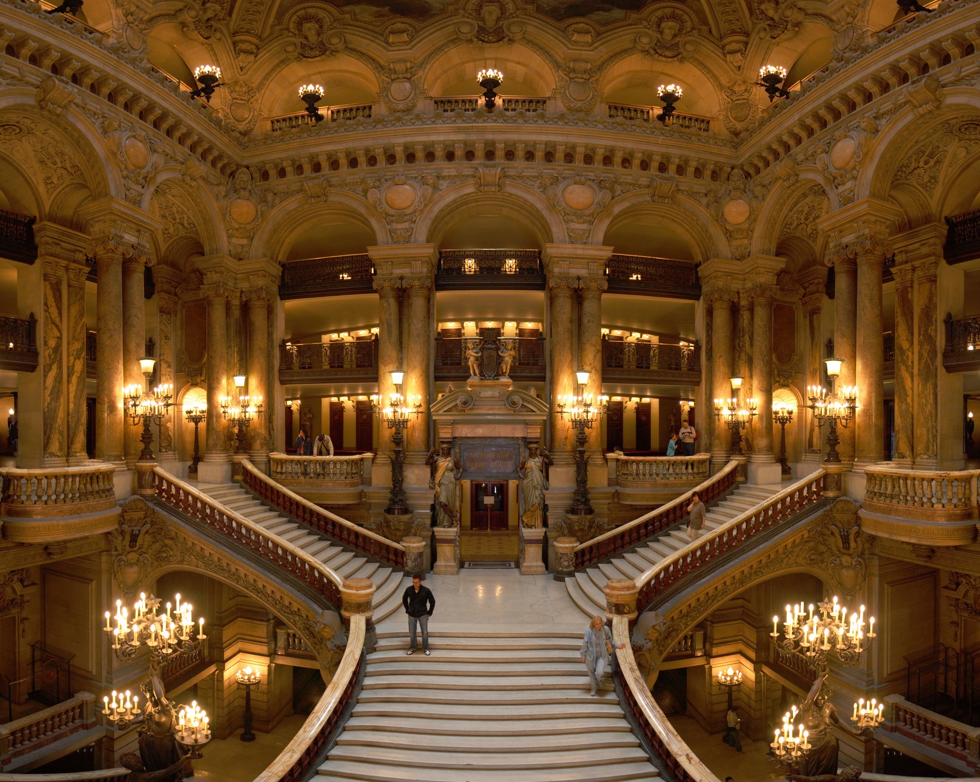 il gran scalone del palais garnier visto dal basso, con le doppie rampe di marmo bianco che si incrociano, la balaustra scura, i lampadari a candelabro accesi e le arcate dorate del livello superiore