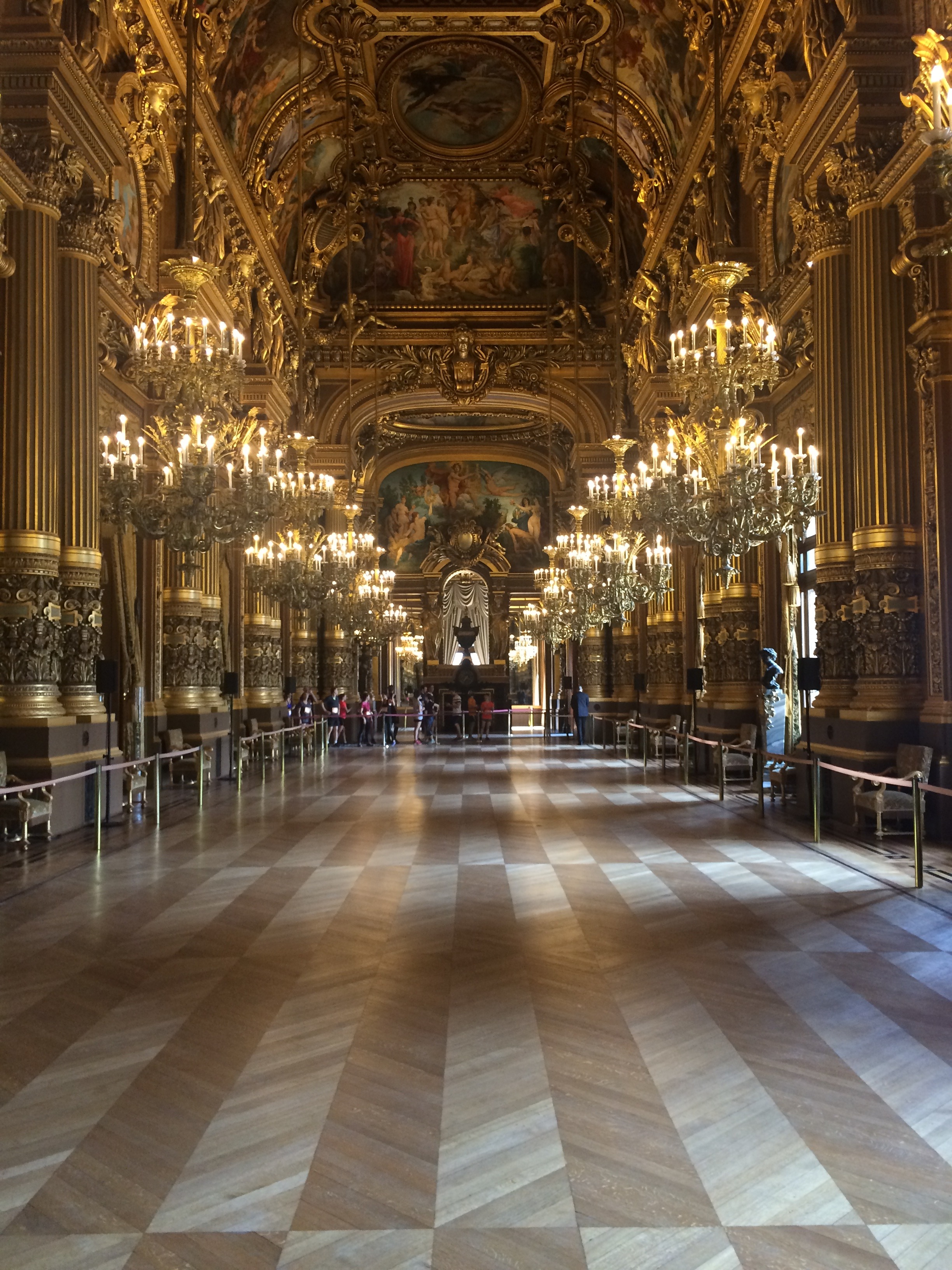 la galleria del grand foyer del palais garnier con le colonne dorate, i lampadari a più bracci accesi in fila, il soffitto affrescato da paul baudry e il pavimento a intarsio geometrico