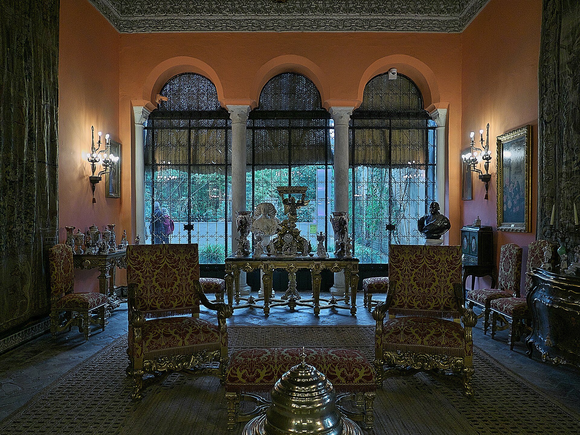 il salon de la gitana del palacio de las duenas, con poltrone dorate, un tavolo con statuette e tre finestre ad arco con le grate aperte sul giardino