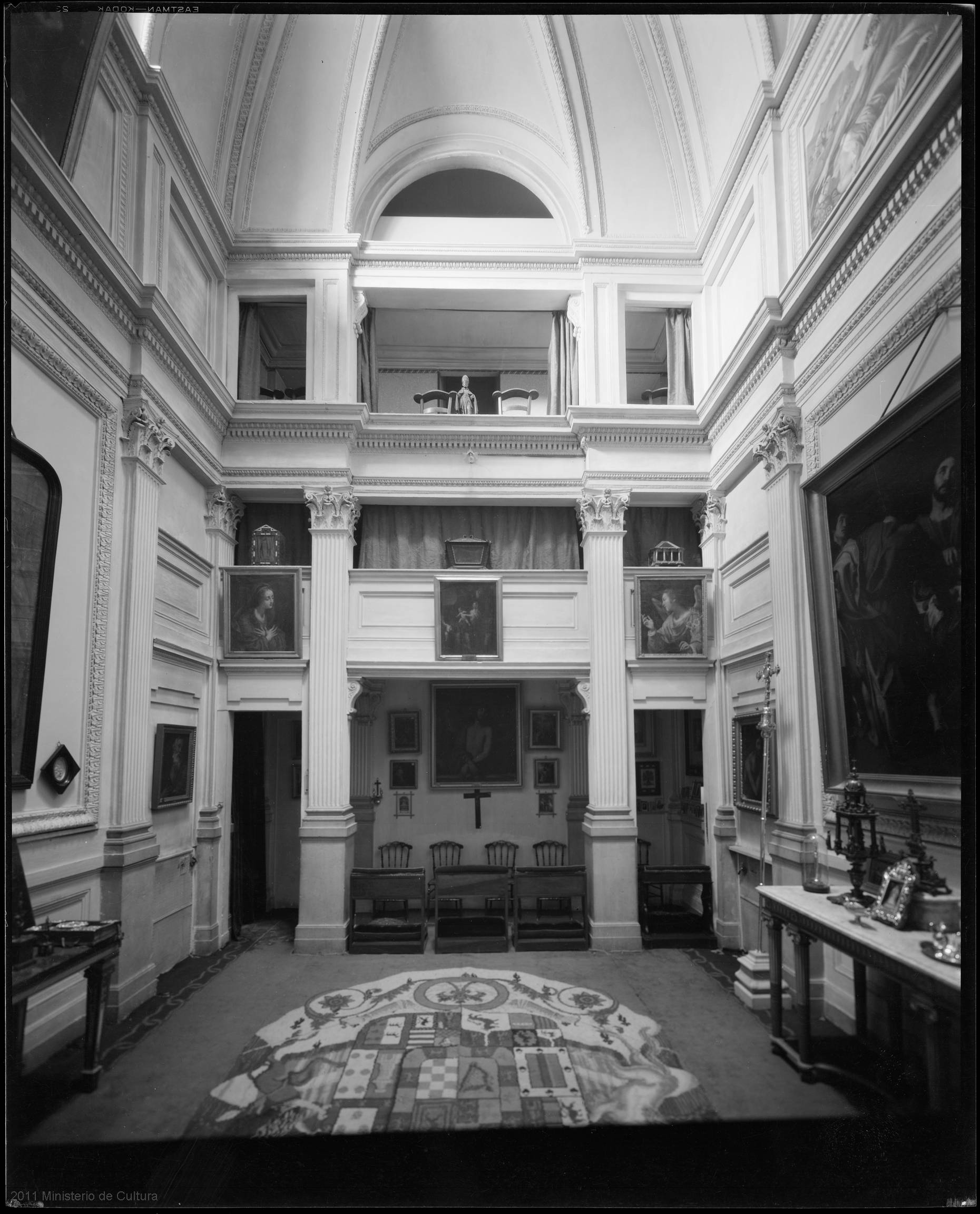 la cappella del palacio de liria fotografata nel 1934, in bianco e nero, con un crocifisso alla parete, le sedie e gli inginocchiatoi, i quadri alle pareti e un tappeto con uno stemma