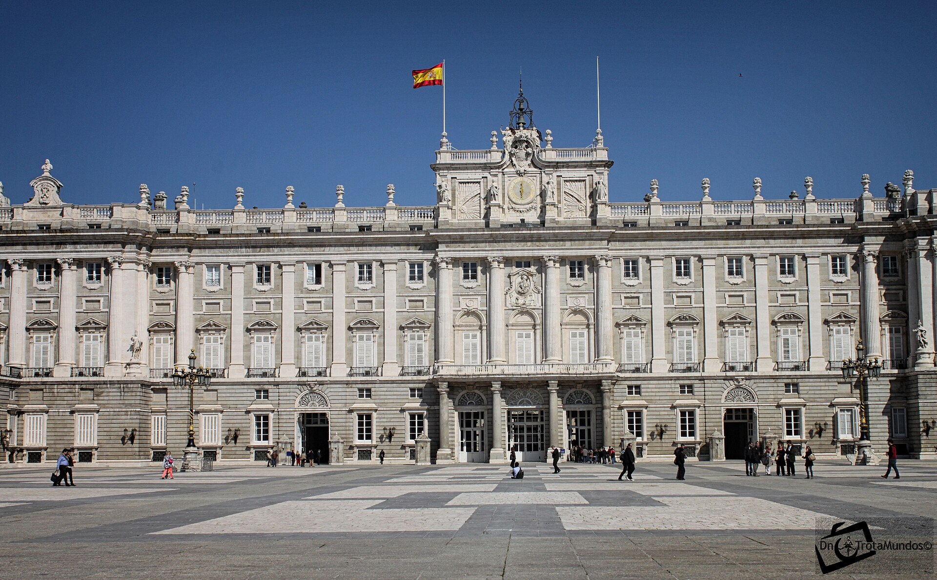 la facciata del palacio real di madrid sulla plaza de la armeria, in pietra chiara, con le colonne, l’orologio e la bandiera spagnola al centro e la grande piazza lastricata davanti