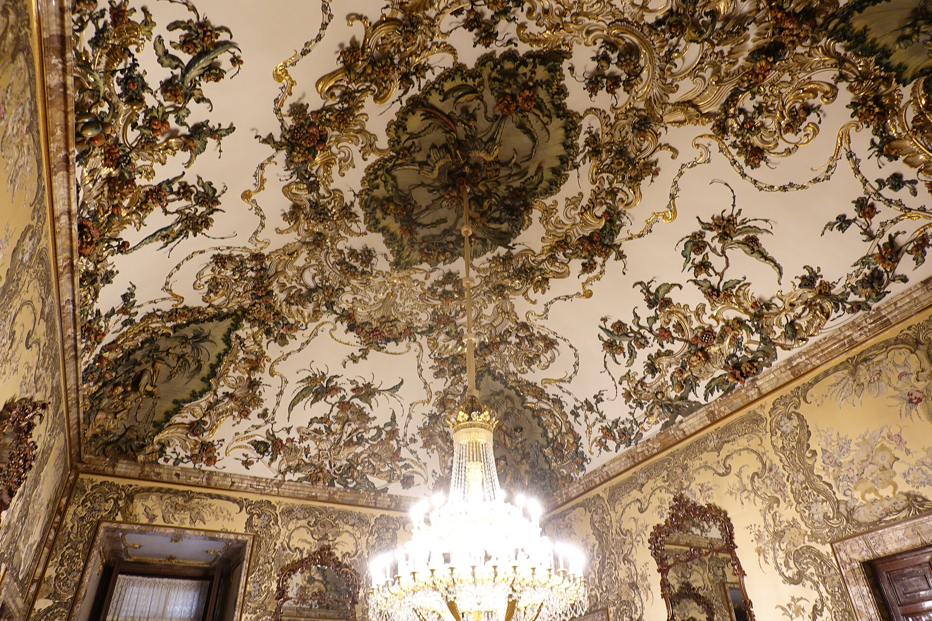 il soffitto della sala di gasparini nel palacio real di madrid, coperto di rami, foglie e fiori in rilievo su fondo chiaro, con un grande lampadario di cristallo