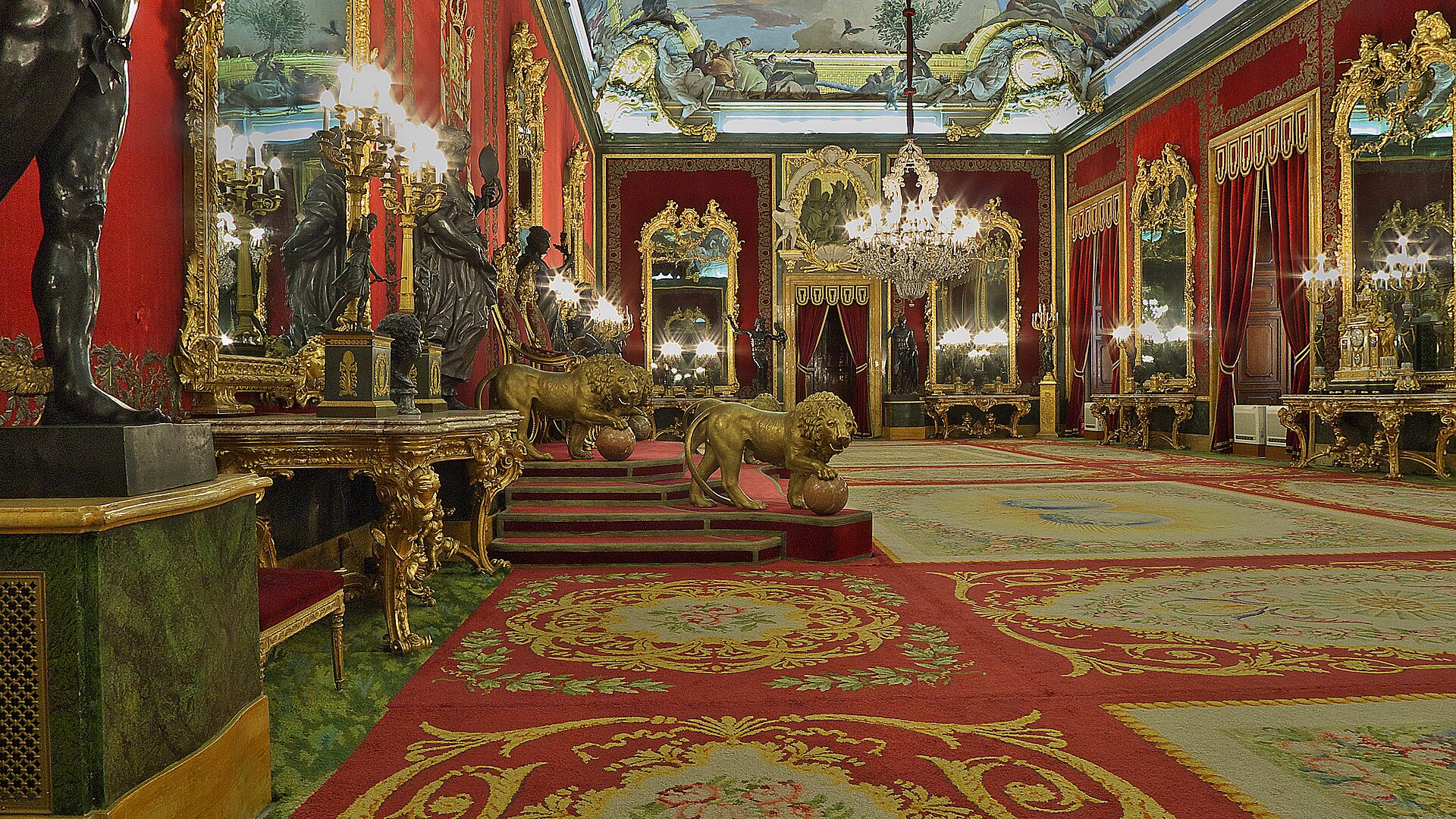 la sala del trono del palacio real di madrid, con le pareti di velluto rosso, gli specchi dorati, il lampadario di cristallo, i leoni di bronzo dorato sui gradini del trono, le statue di bronzo e il tappeto rosso