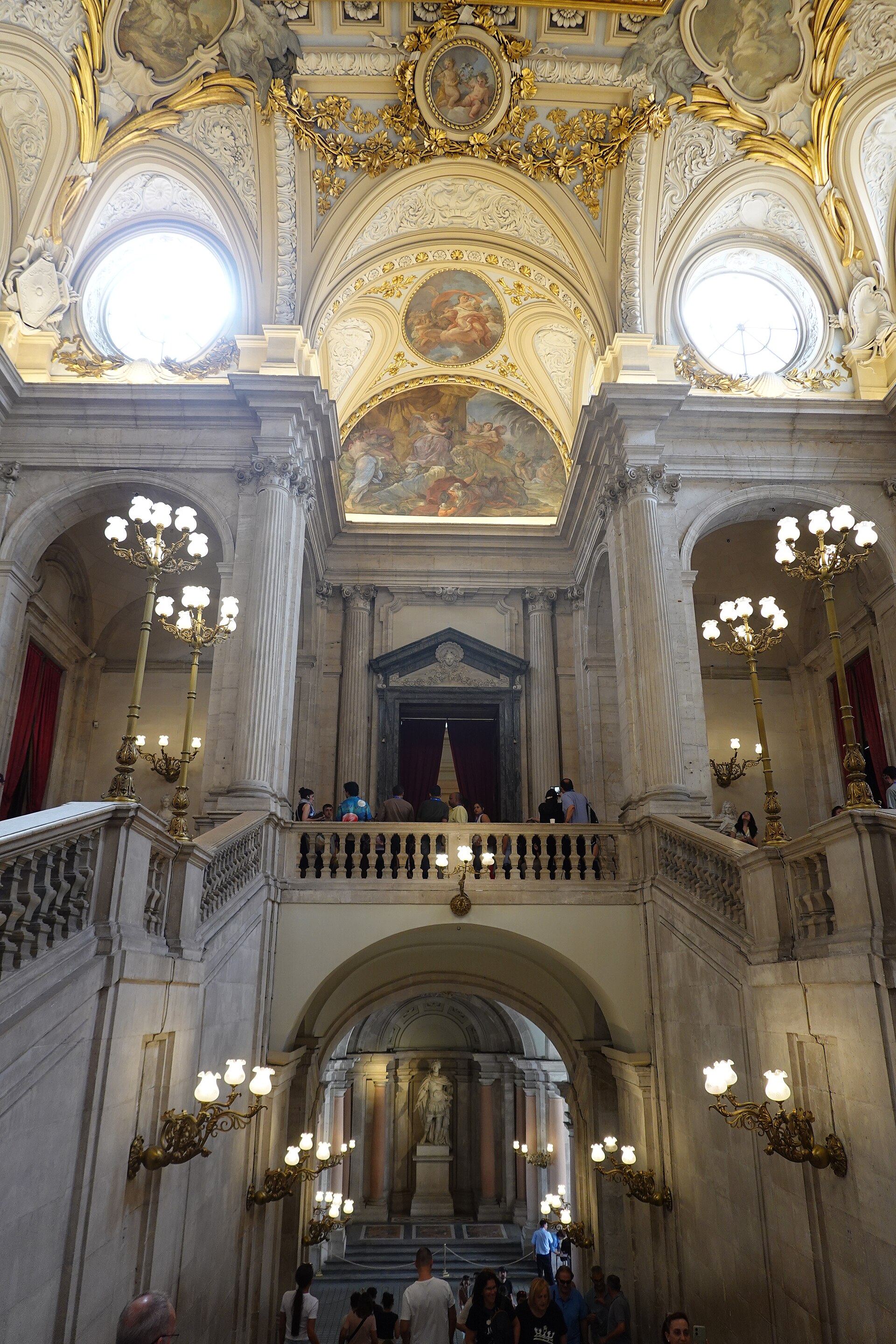 lo scalone principale del palacio real di madrid visto dal basso, con le balaustre del pianerottolo, i candelabri dorati, la volta affrescata con le grandi finestre rotonde e, sotto l’arco, una statua