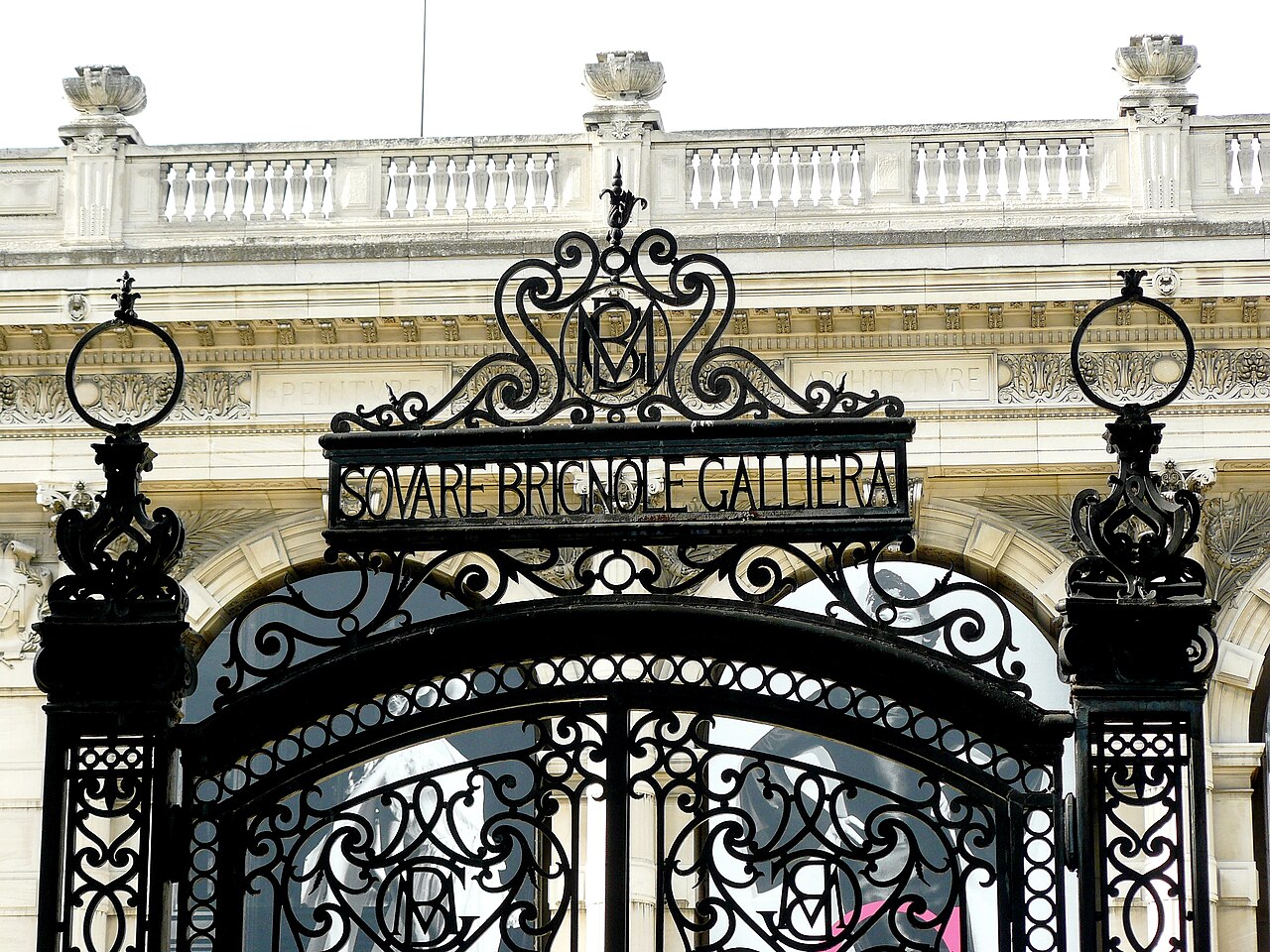il cancello in ferro battuto del square brignole galliera con il monogramma della duchessa e il fregio del palazzo con la parola peinture sullo sfondo