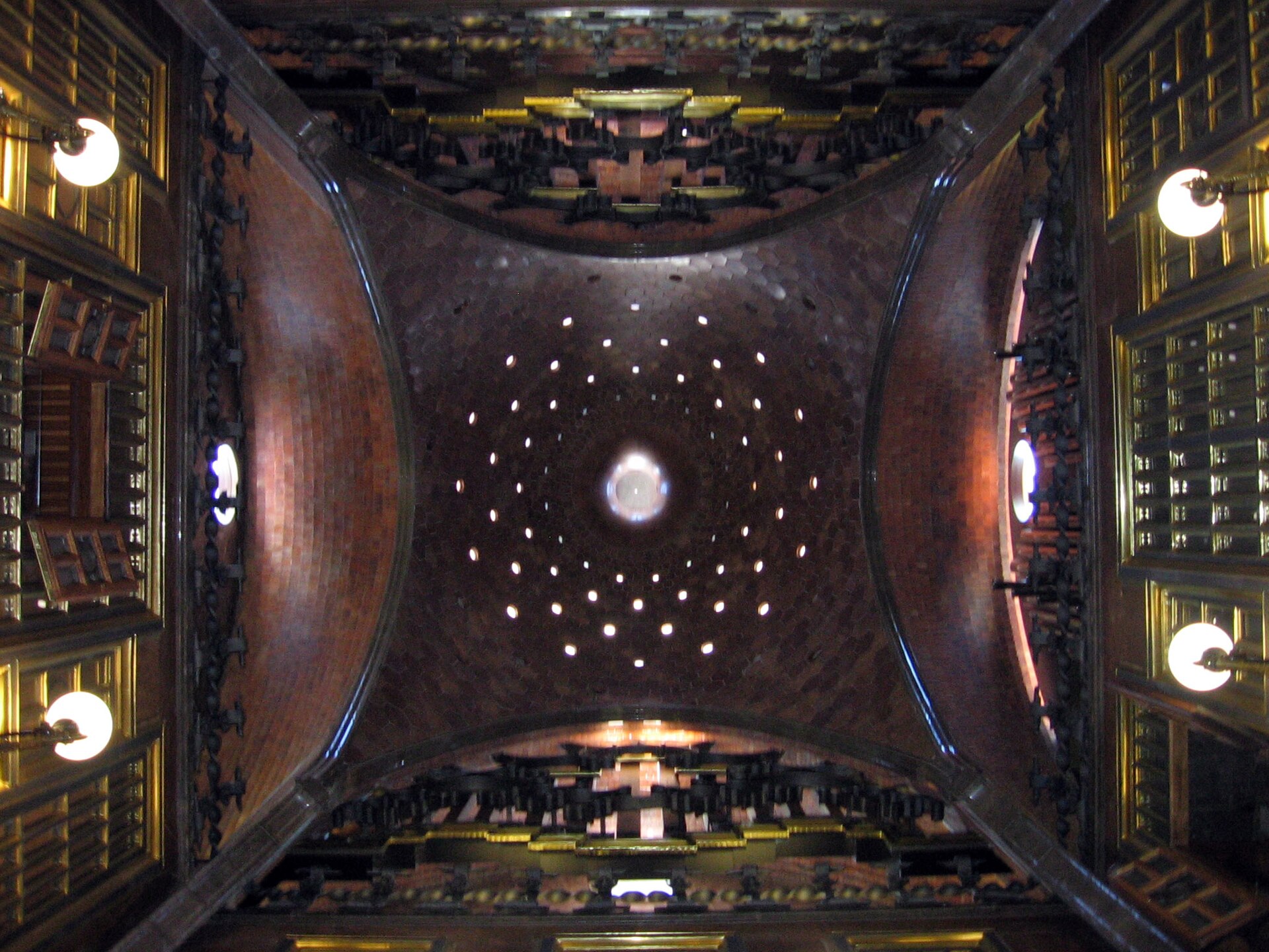 la cupola del salone del palau guell vista dal basso, scura e forata da cerchi di piccoli fori luminosi attorno a un’apertura centrale