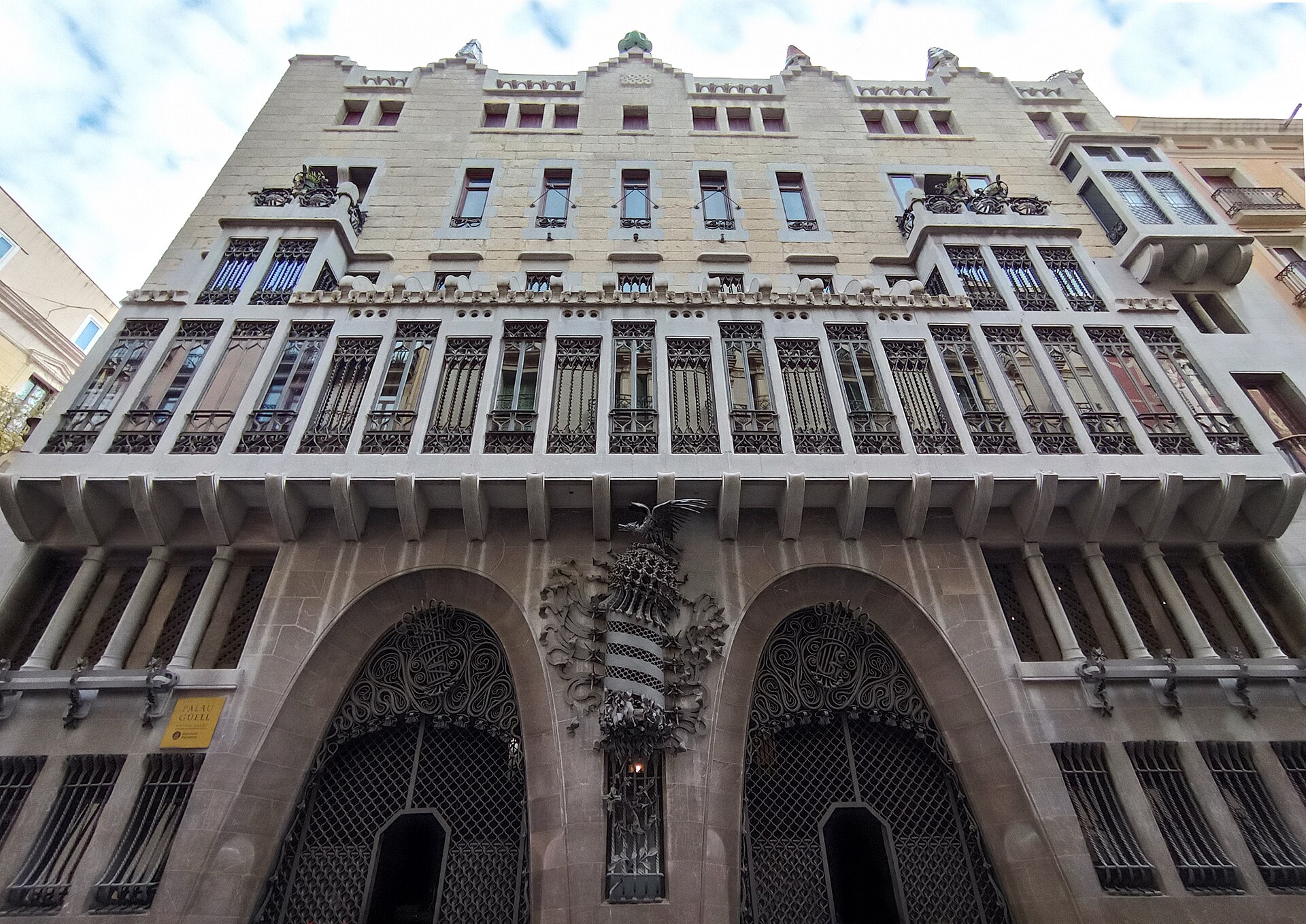 la facciata di pietra del palau guell vista dal basso, con i due portoni ad arco parabolico, lo stemma con la fenice fra i due e la tribuna sopra