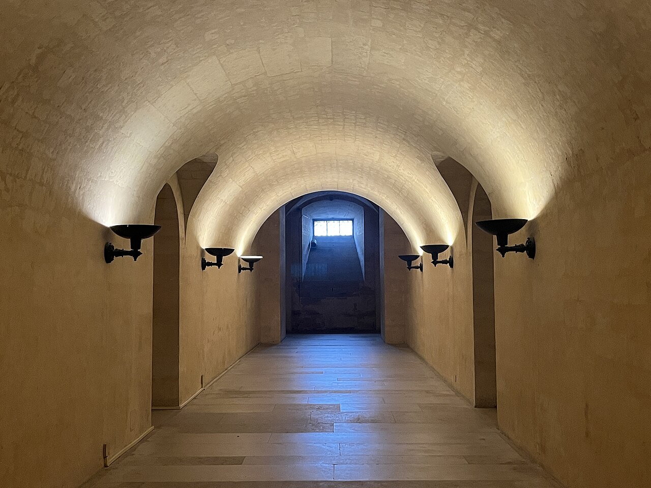 un corridoio della cripta con la volta a botte in pietra chiara, le applique nere a coppa che illuminano la volta dal basso su tutti e due i lati, il pavimento in lastre di pietra e in fondo una finestrella sbarrata