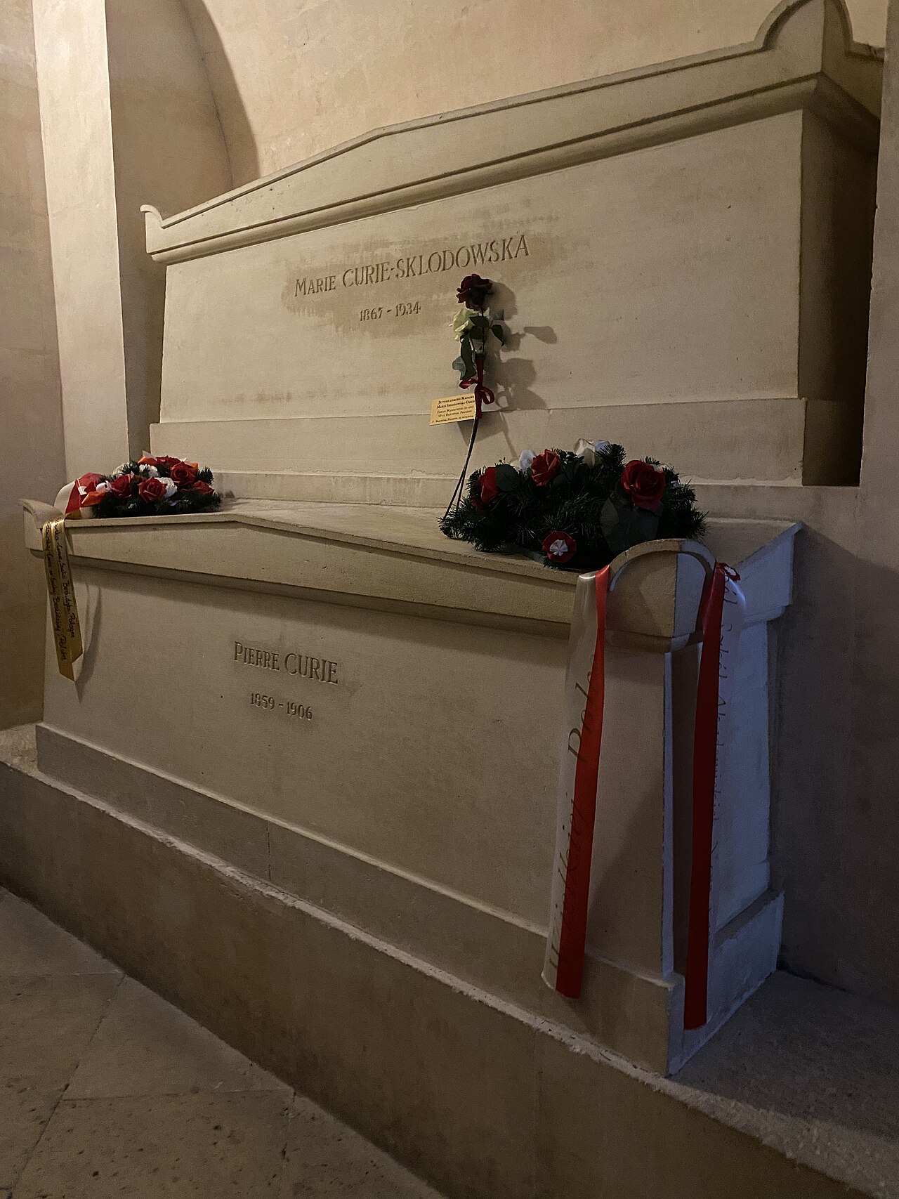 le due tombe di pietra chiara sovrapposte nella cripta, quella di marie curie sklodowska 1867 1934 in alto e quella di pierre curie 1859 1906 in basso, con corone di fiori rossi e bianchi e nastri appoggiati sul bordo