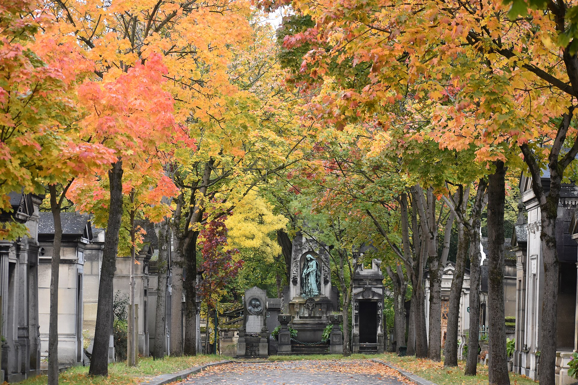 un viale del cimitero del pere lachaise a parigi in autunno, con gli aceri gialli e arancioni sopra le cappelle di pietra e una statua di bronzo verde in fondo al viale