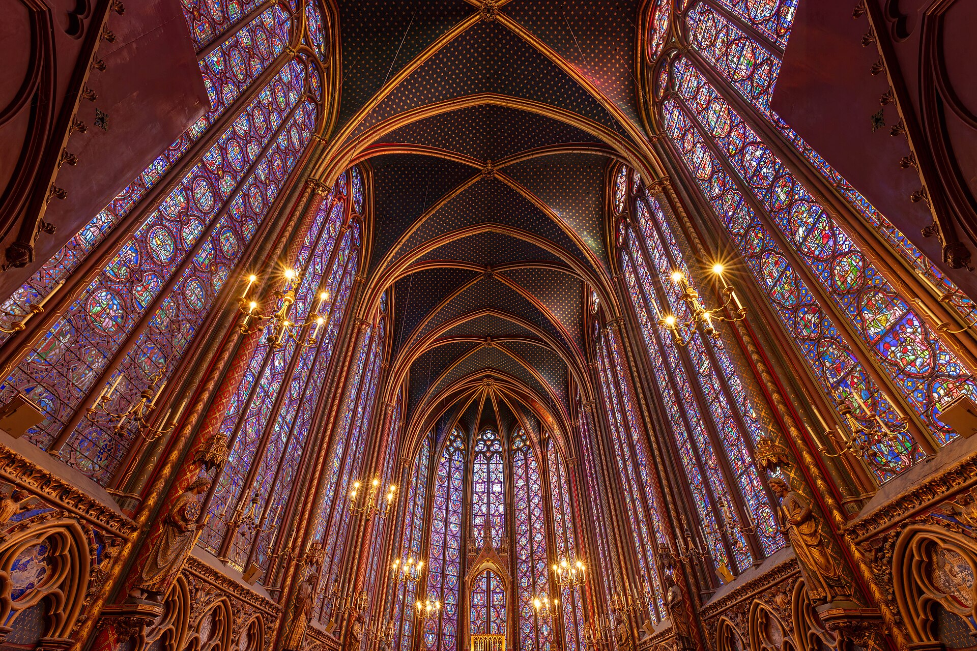 la navata alta della sainte chapelle di parigi vista dal basso, con le vetrate colorate che salgono fino alla volta blu punteggiata di stelle dorate e i lampadari accesi lungo le pareti