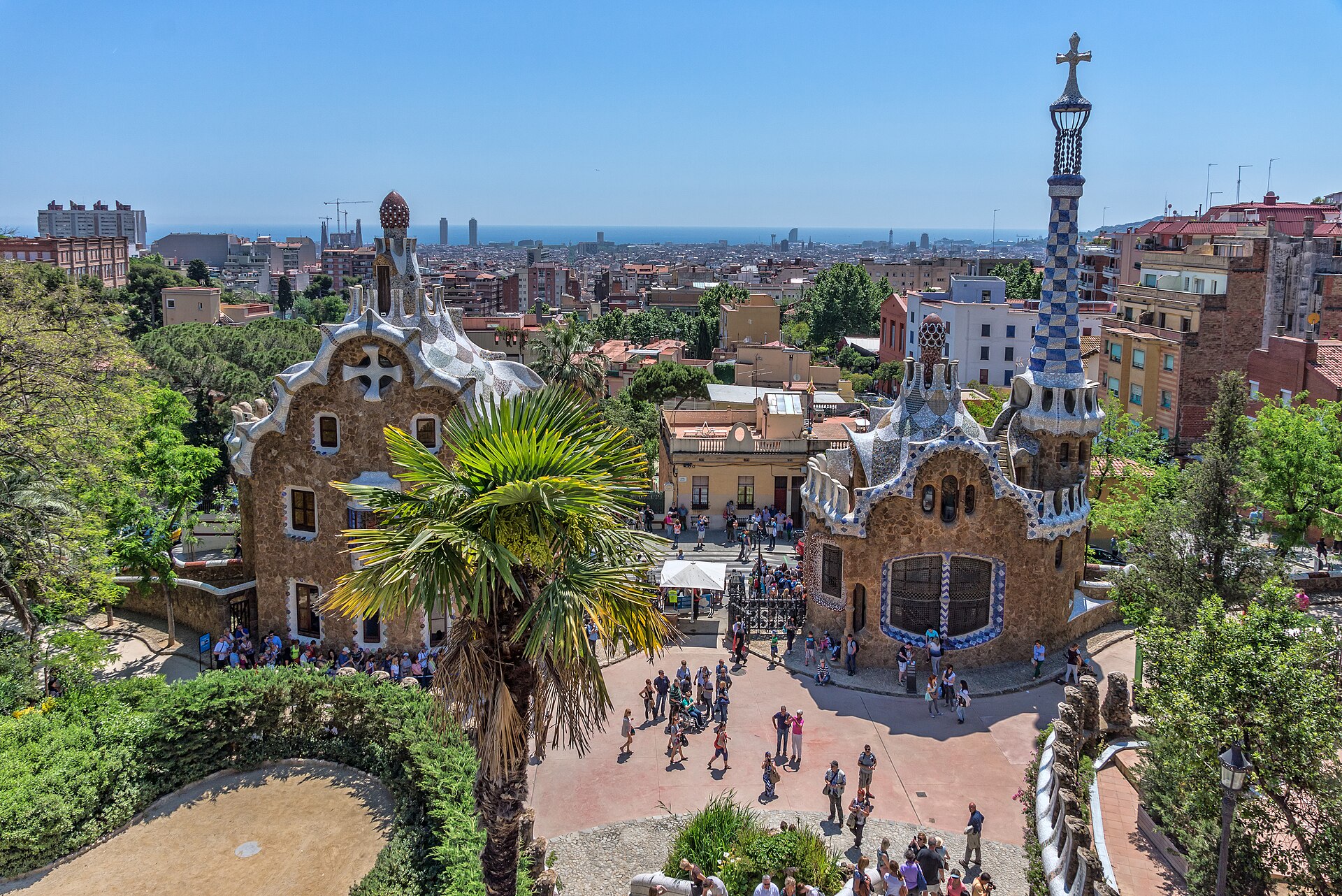 i due padiglioni d’ingresso di park guell visti dall’alto, con i tetti a mosaico, la torre a scacchi e la città sullo sfondo
