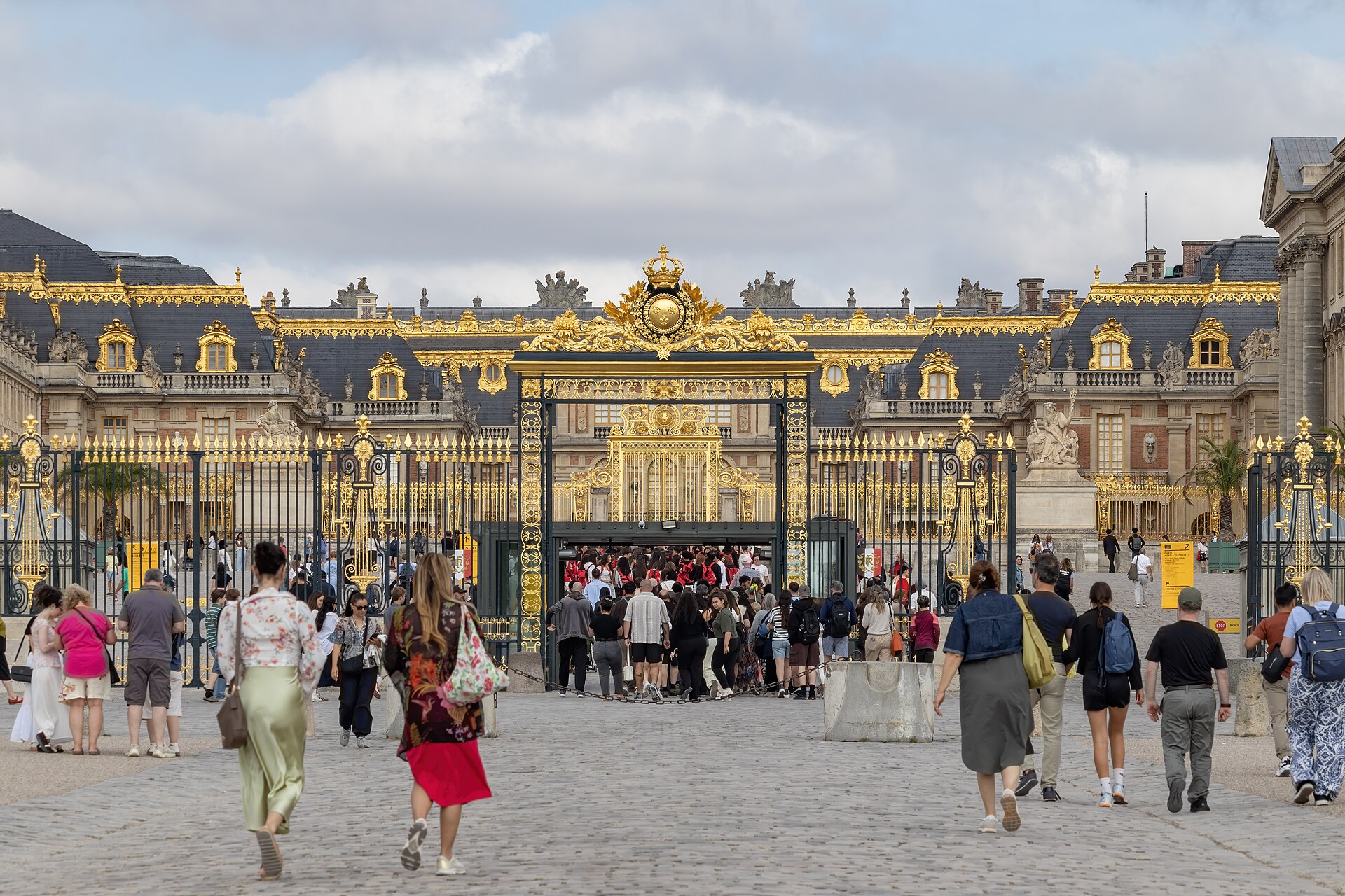 il cancello dorato della reggia di versailles visto dal cortile, con il tetto in ardesia e gli ornamenti in oro zecchino sulla facciata e i visitatori che si avviano verso l’ingresso