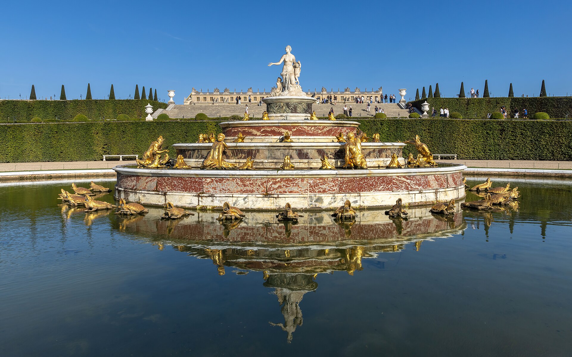 la fontana di latona nei giardini di versailles, con le statue dorate delle rane intorno alla vasca a gradoni e il palazzo sullo sfondo oltre il prato