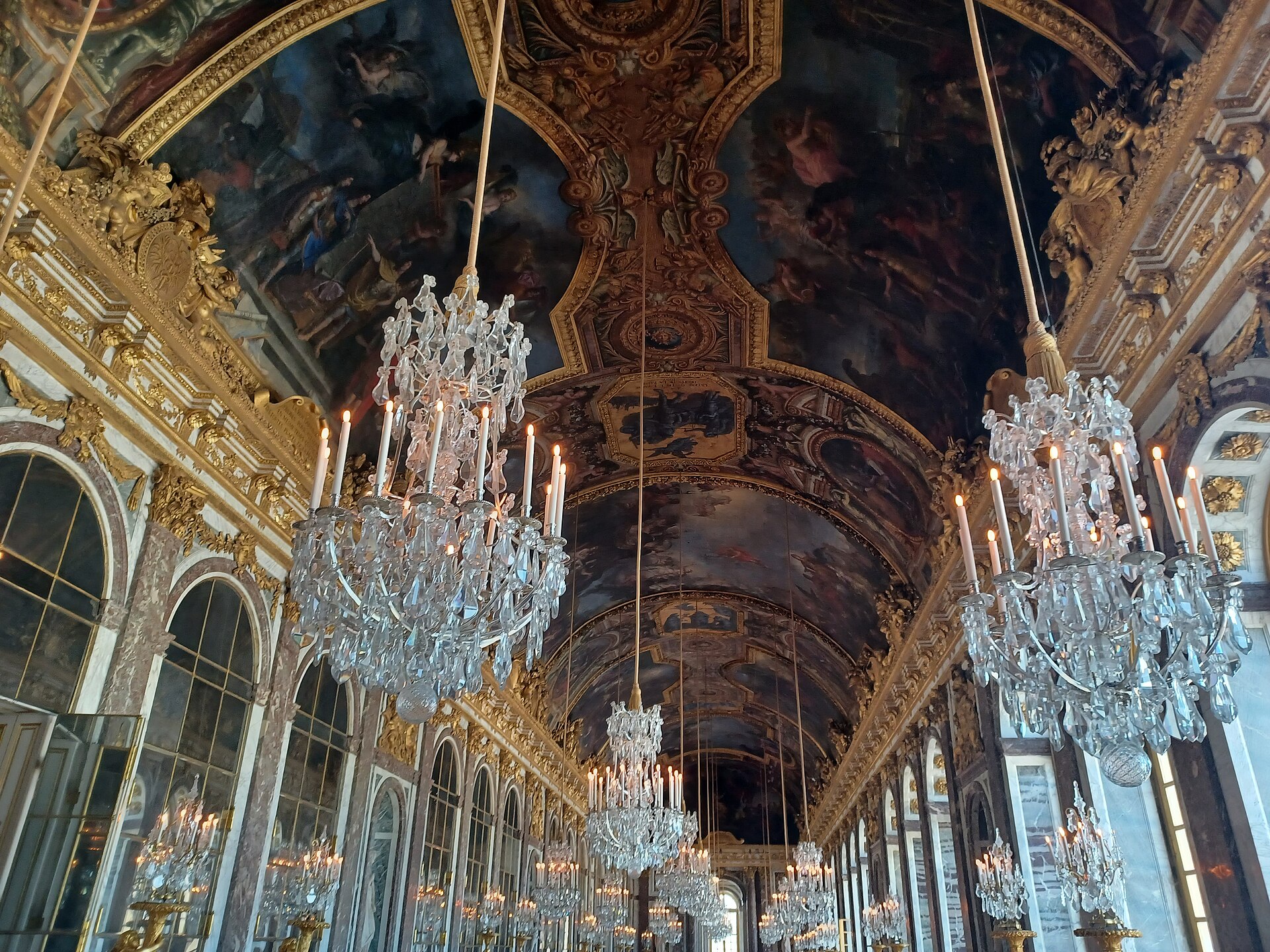 la volta dipinta della galleria degli specchi di versailles vista dal basso, con due lampadari di cristallo accesi in primo piano e altri lampadari in fila lungo la galleria, in una fotografia del 2023 precedente al nuovo allestimento