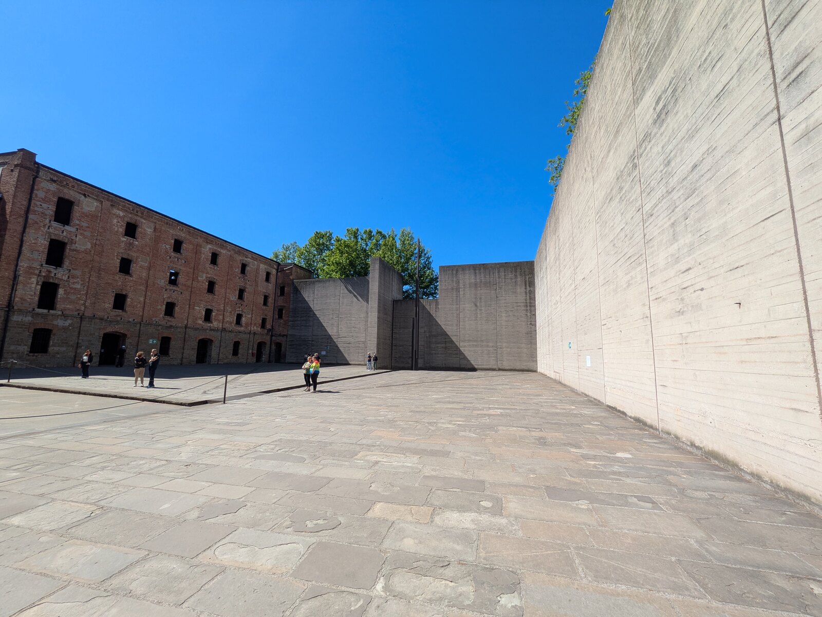 il muro di cemento a vista che recinta il cortile della risiera di san sabba accanto agli edifici in mattoni
