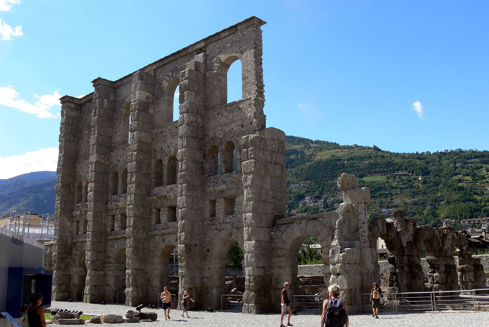 il teatro romano di aosta visto da ovest, con la facciata e i piloni della scena sotto il cielo azzurro e la montagna dietro