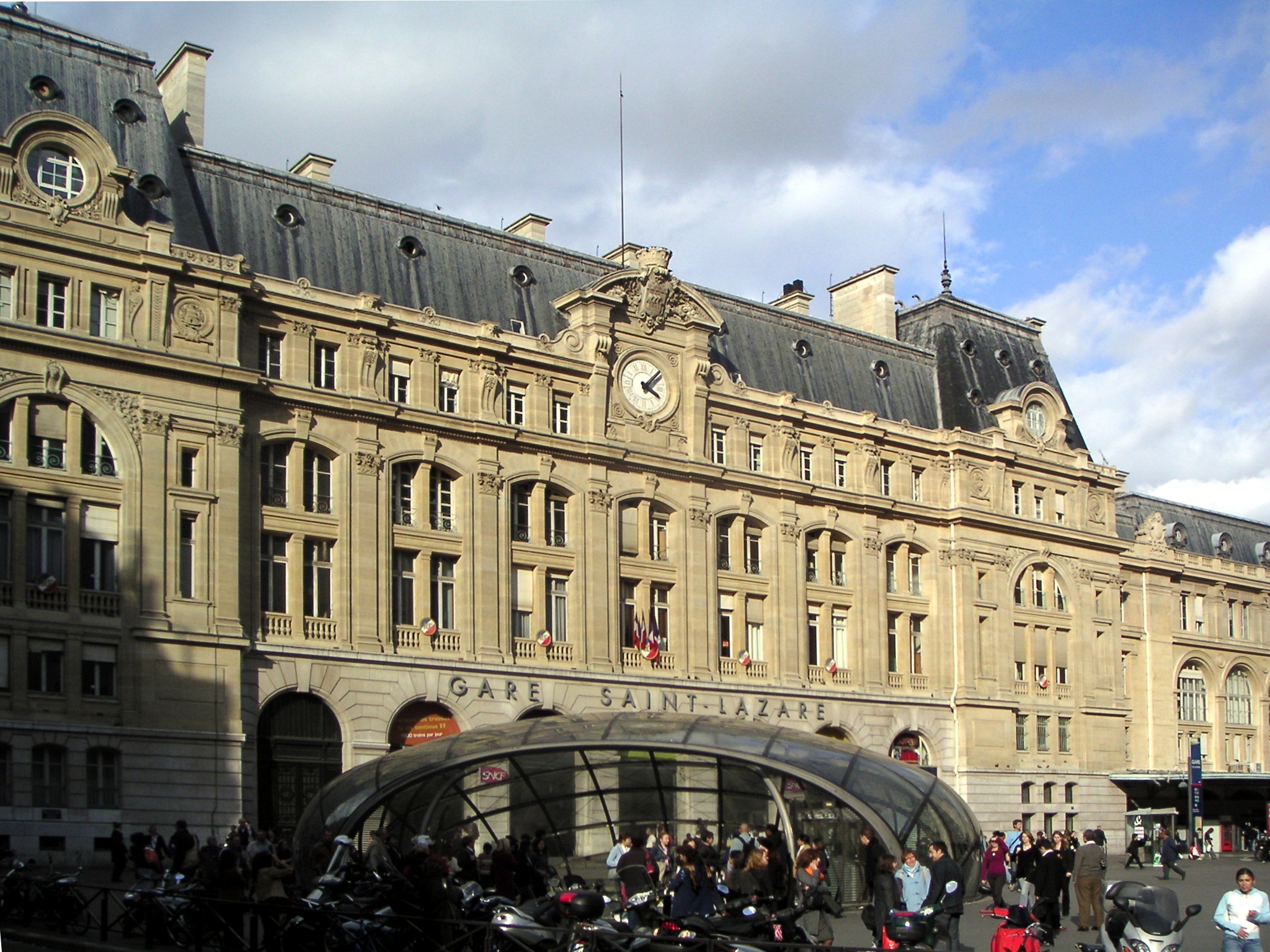 la facciata della gare saint-lazare a parigi con l’orologio centrale sopra l’ingresso della metropolitana e i passanti davanti alla pensilina in vetro