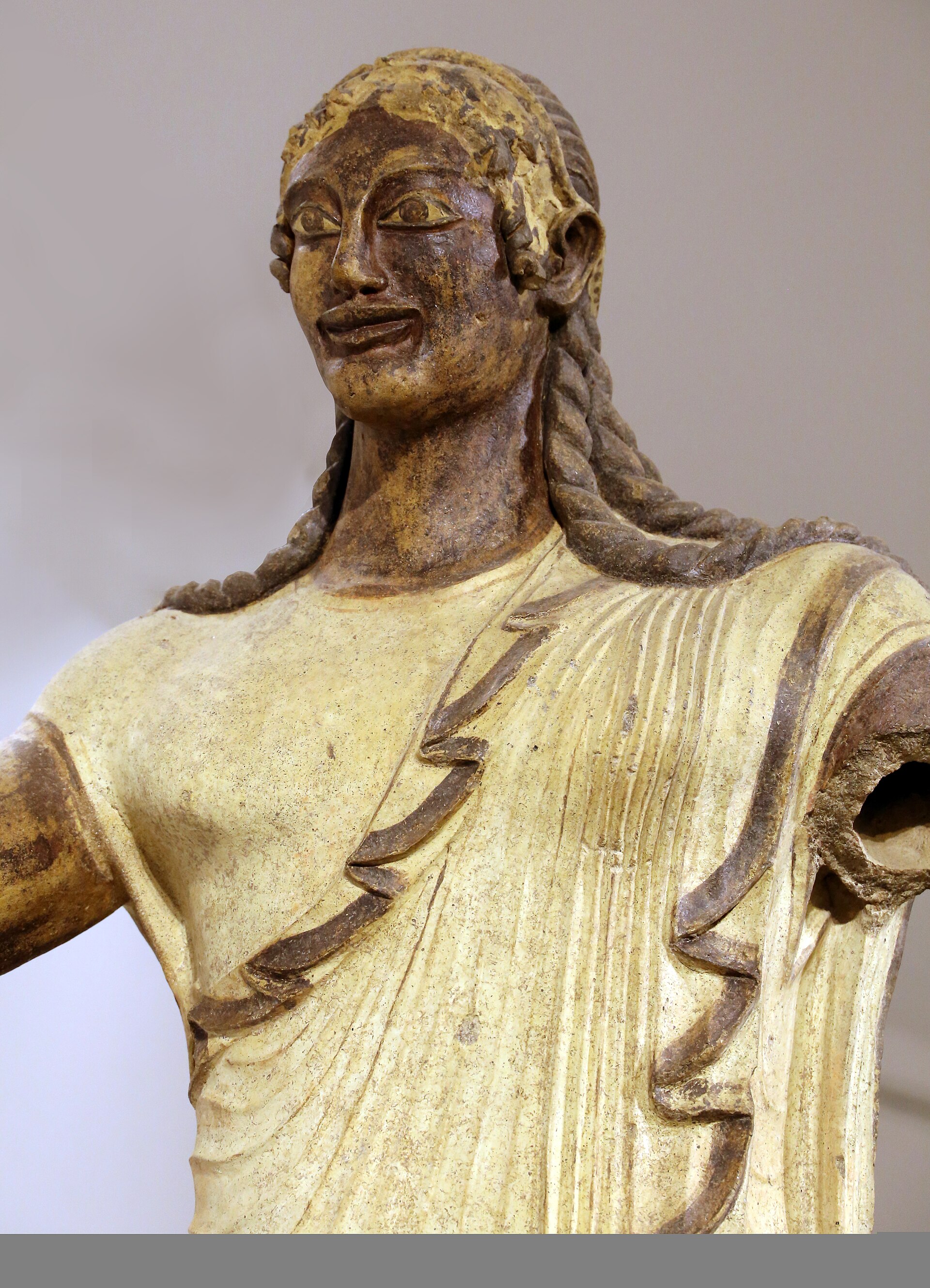 il busto dell apollo di veio in terracotta, con il sorriso largo, le trecce sulle spalle e il mantello inciso a solchi profondi