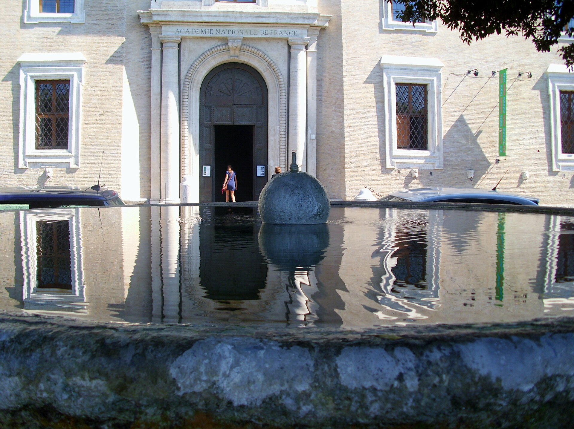 the fountain in front of the entrance of villa medici, with the dark stone sphere in the middle of the round basin and the arched doorway with the inscription of the french academy reflected in the water