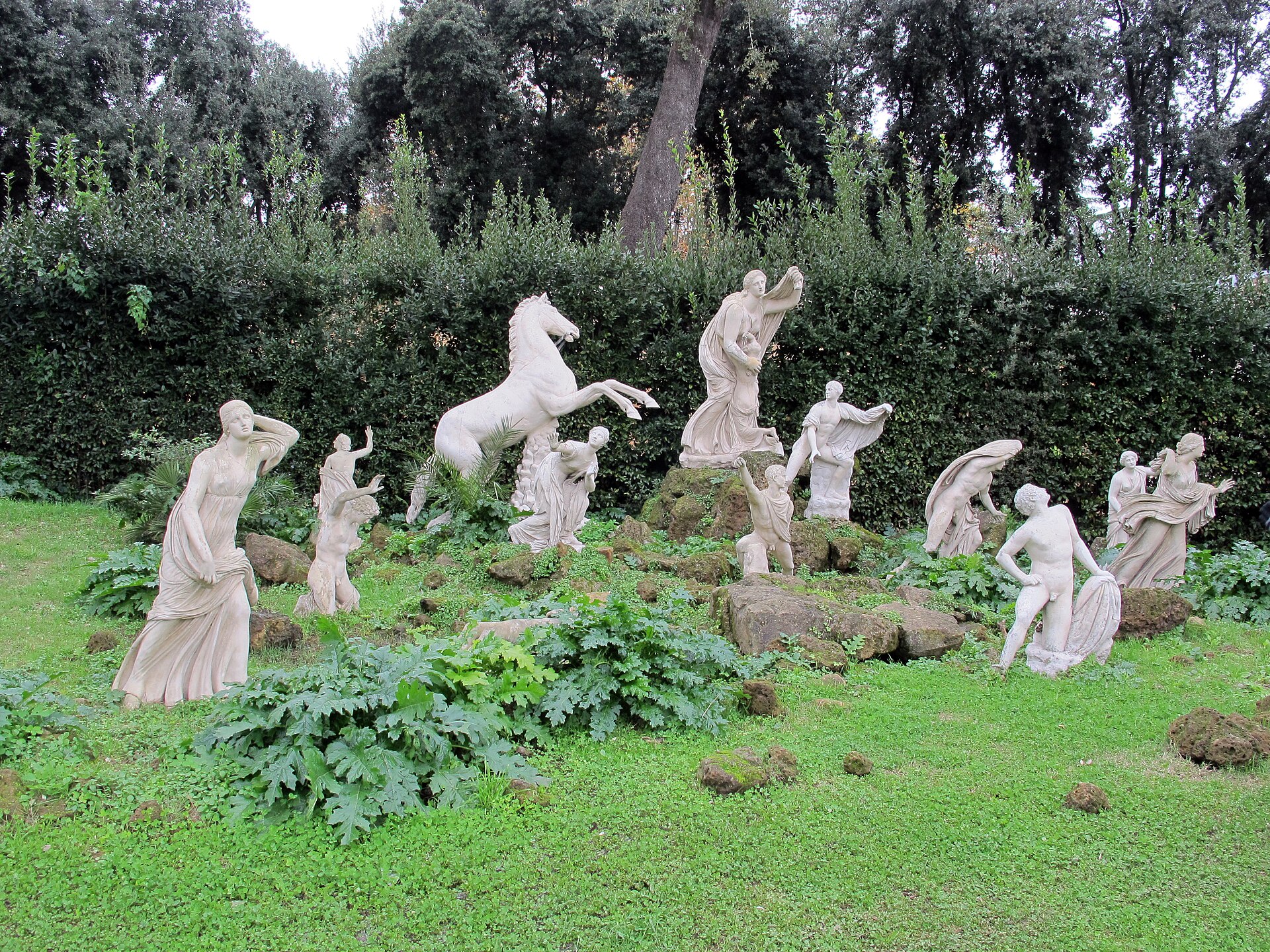 the white marble copies of the niobids spread over a lawn with rocks, among them a rearing horse and figures in motion, with a tall hedge and the holm oaks of the garden in the background