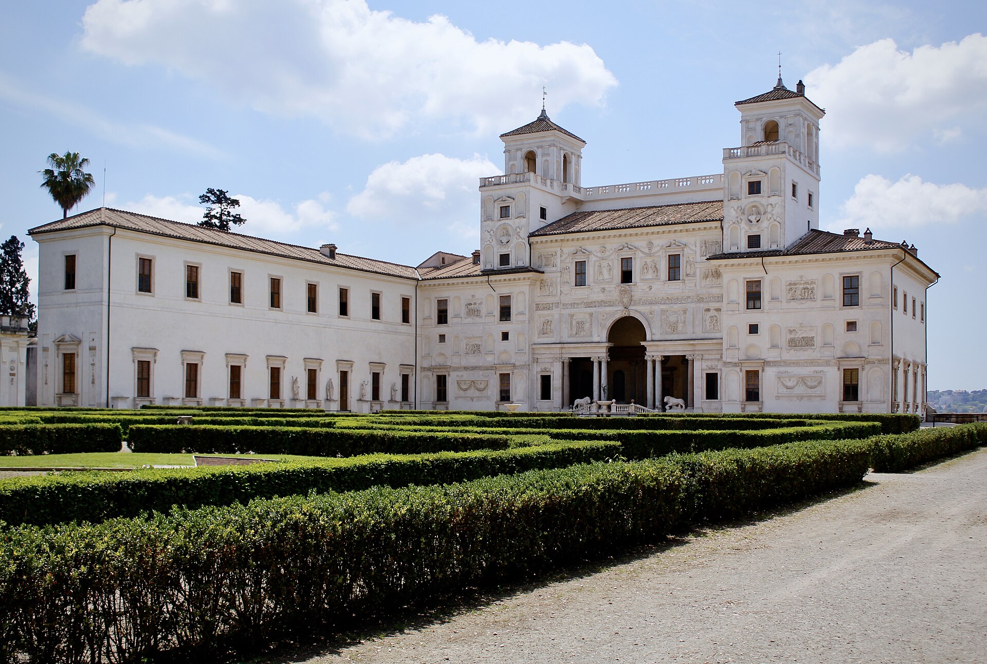 the garden front of villa medici, white and covered in ancient reliefs, with the two towers, the colonnaded loggia in the centre with two lions beside the steps and clipped box hedges in the foreground